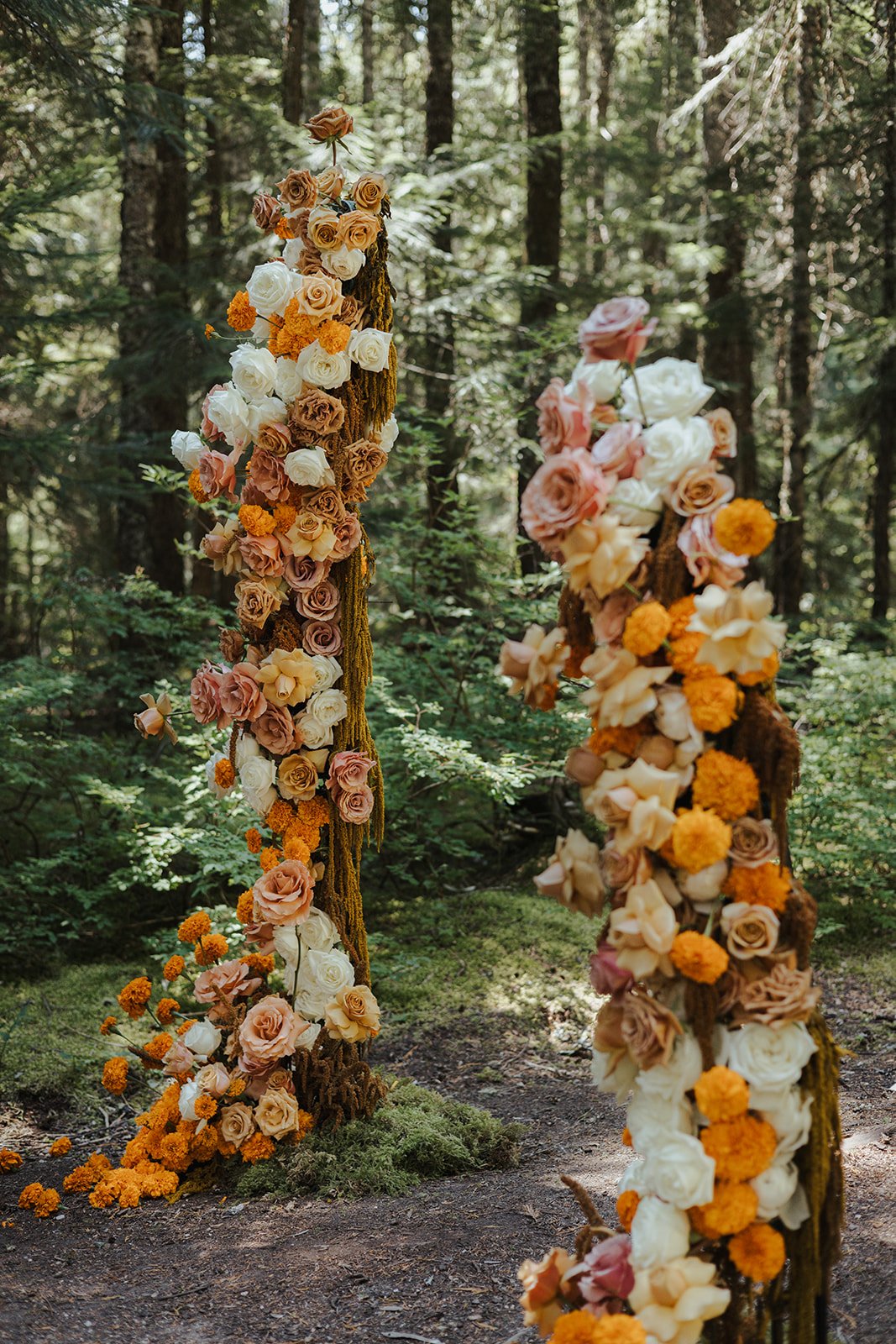 Two wooden posts wrapped with flowers including roses in shades of peach, pink, cream, and white, as well as orange marigolds, in a forest setting.