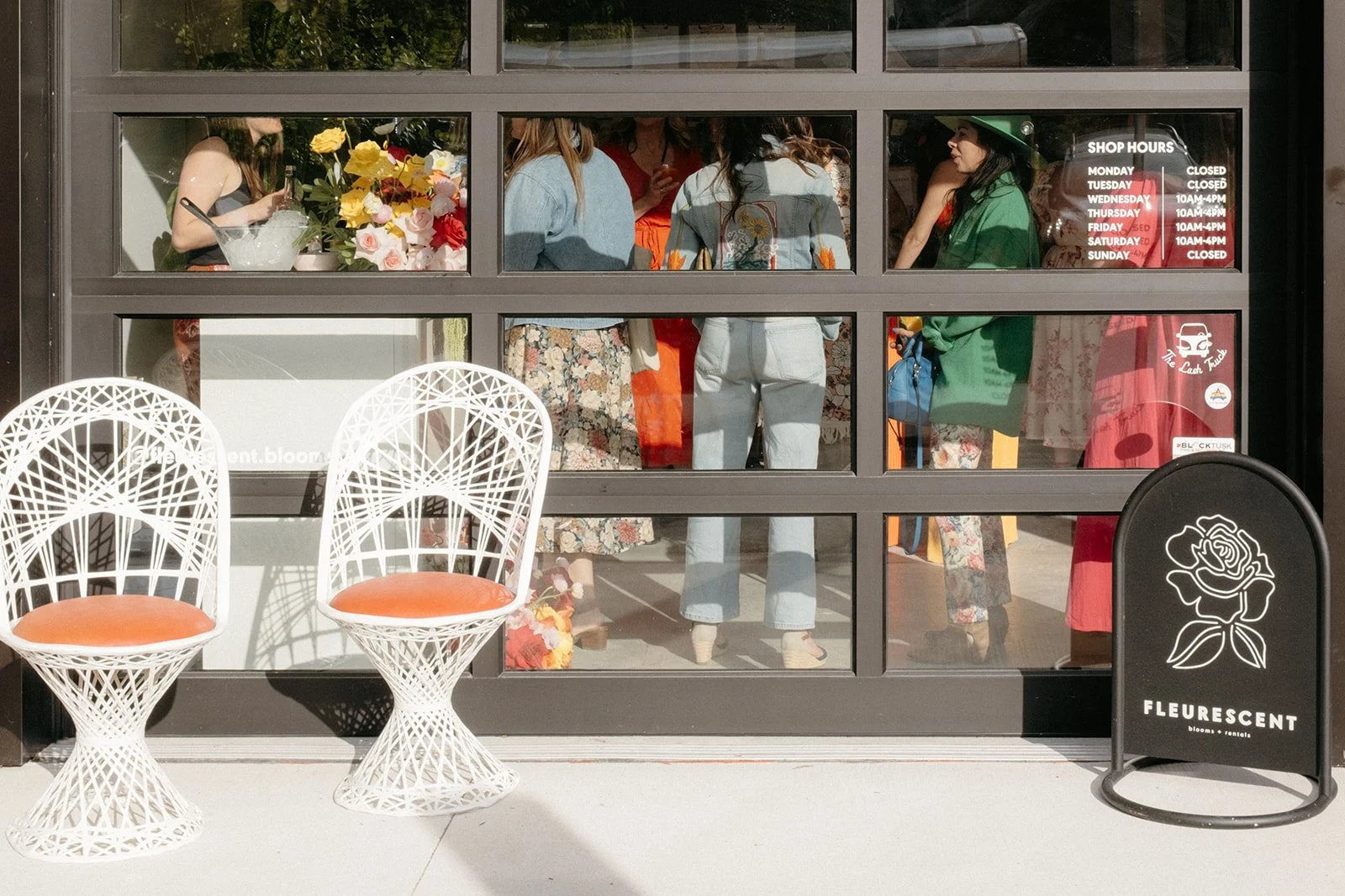 People inside a flower shop viewed through large glass windows, with two white decorative chairs with orange cushions outside, and a black sign with a rose illustration and the word 'FLEURSÉNT'.