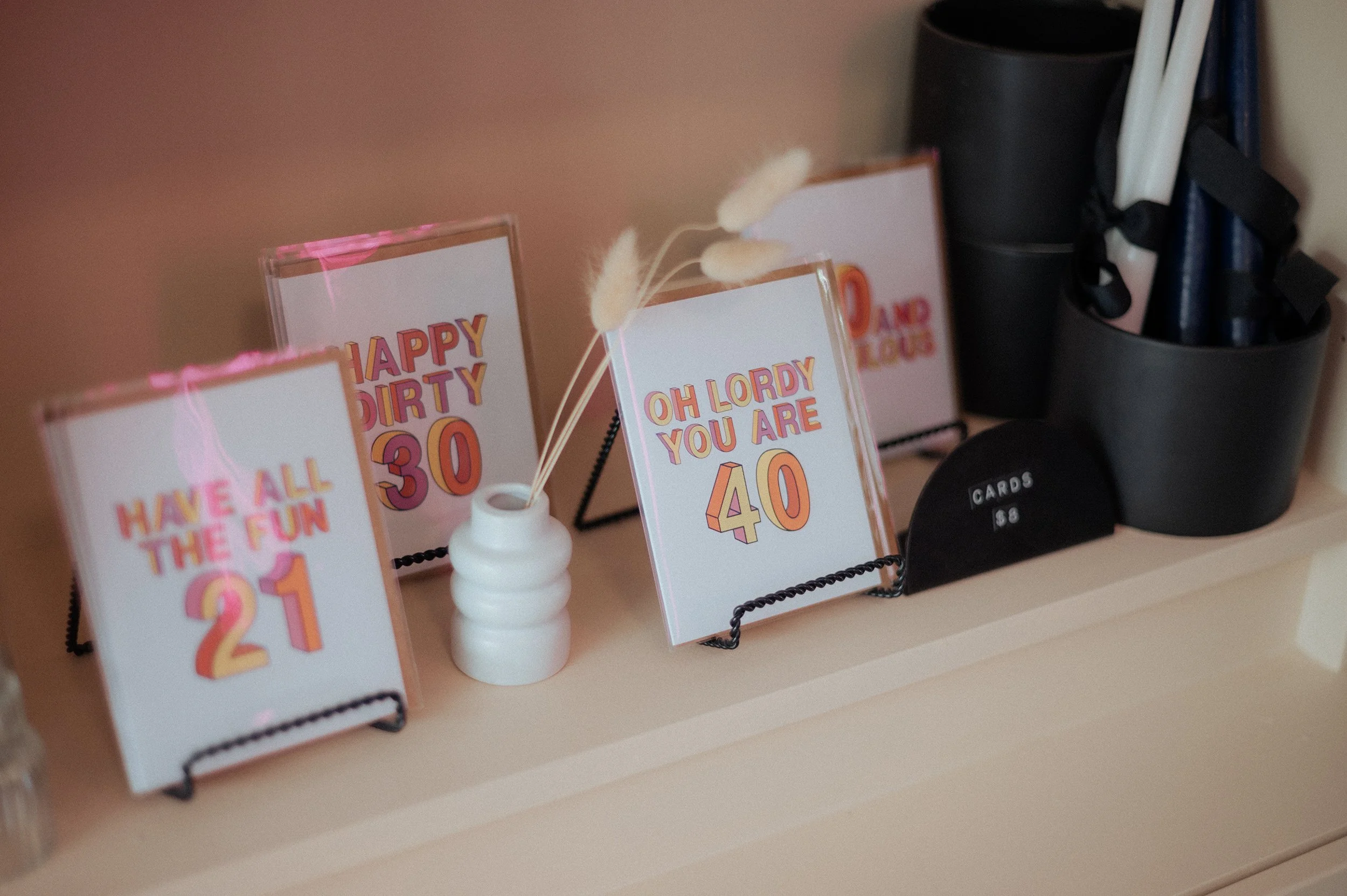 Decorative signs with birthday messages and ages, including 'HAVE ALL THE FUN 21,' 'HAPPY DIRTY 30,' 'OH LORDY YOU ARE 40,' arranged on a black metal stand, with a small white vase holding dried flowers, and black containers with pens or chopsticks i