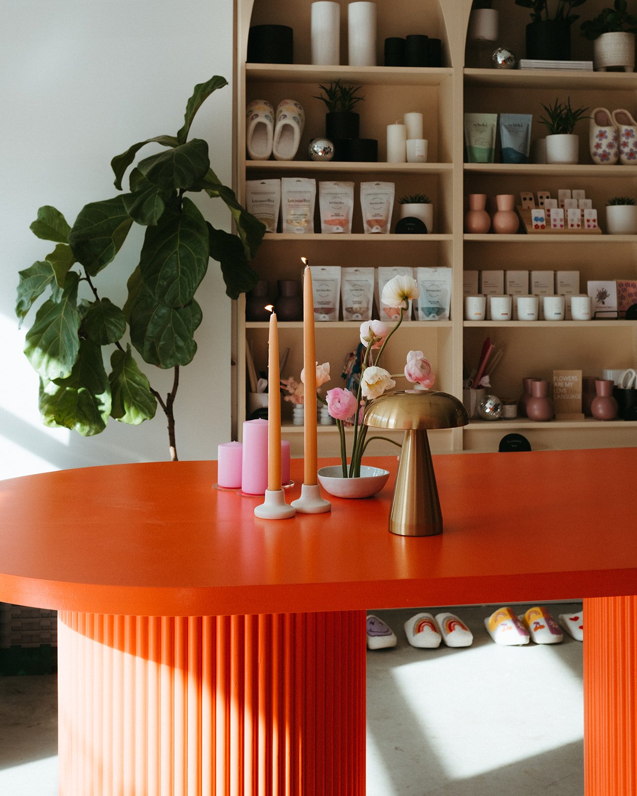 Decorative orange table with pink candles, a white candle holder with two lit candles, a gold vase with pink and white flowers, in a room with a large green leafy plant and a beige shelf filled with candles, small plants, and decorative items.