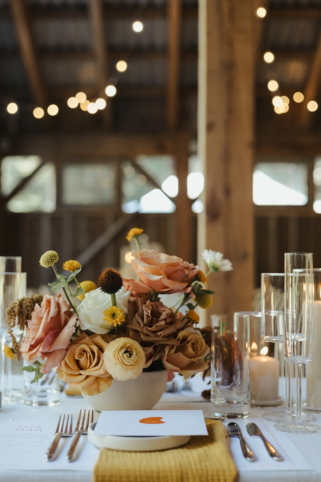 A table setting with a floral centerpiece, candles, and glassware in a rustic barn with string lights overhead.