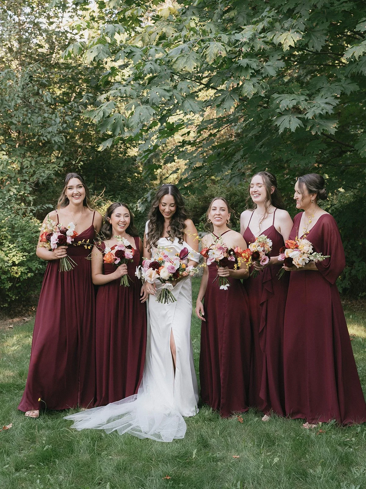 A group of six women, including a bride in a white dress, standing outdoors on grass under a large leafy tree, holding colorful bouquets and smiling.