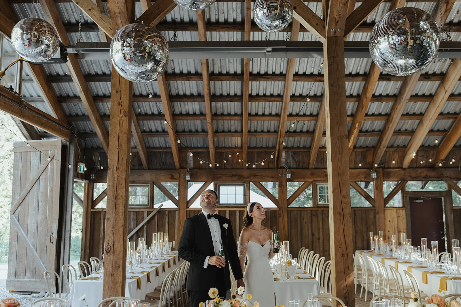 Bride and groom standing inside a rustic wooden barn, smiling and holding drinks, with long decorated tables and disco balls hanging from the ceiling.