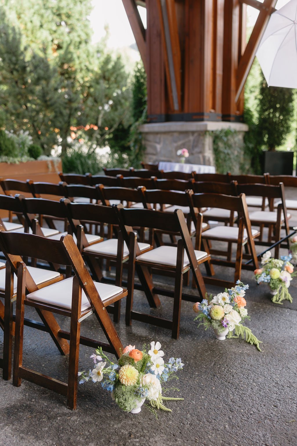 Rows of wooden folding chairs with white cushions arranged for an outdoor wedding ceremony, decorated with flower arrangements in white vases, with greenery and a wooden structure in the background.