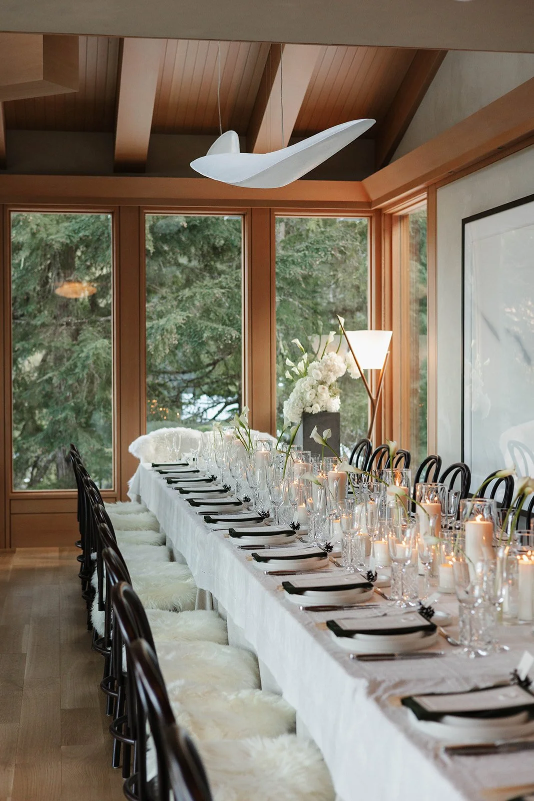 Long dining table set with white tablecloth, black napkins, wine glasses, and candles, in a room with large windows and wooden ceiling, decorated with white flowers and a whale-shaped light fixture.
