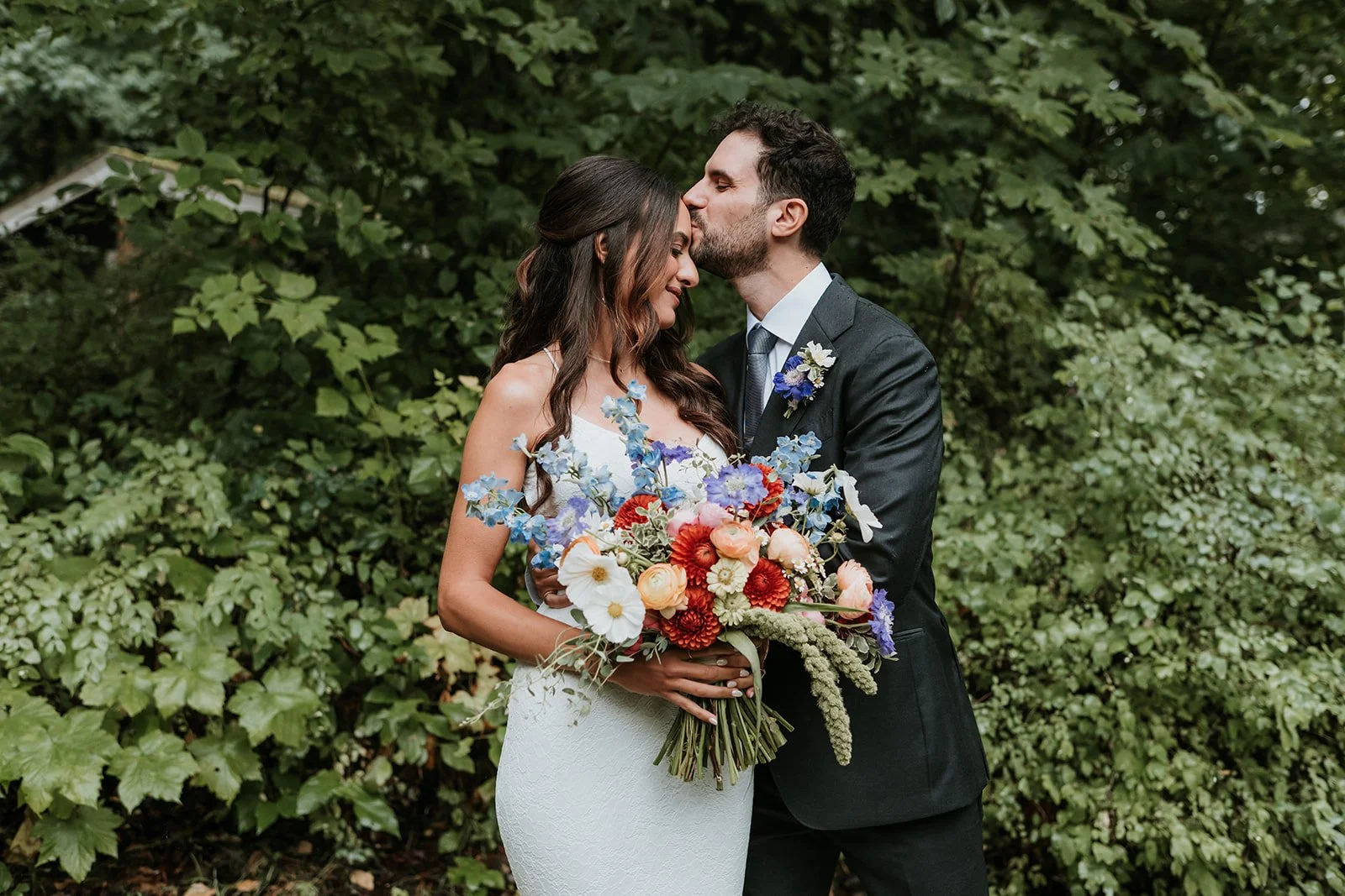 A bride and groom standing close, with the groom kissing the bride's forehead, in front of lush green foliage. The bride is holding a colorful bouquet of flowers.