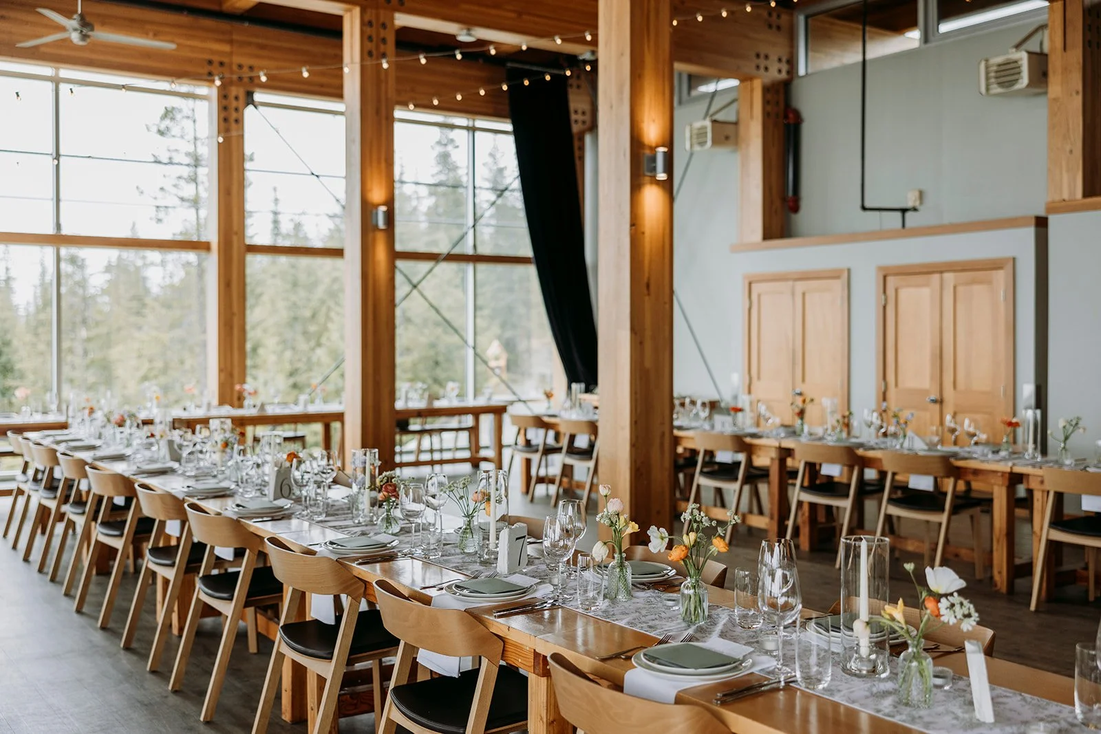 Modern event space with large windows, wooden beams, long rectangular tables set for a banquet, decorated with small vases of flowers, candles, and tableware, natural light coming in.