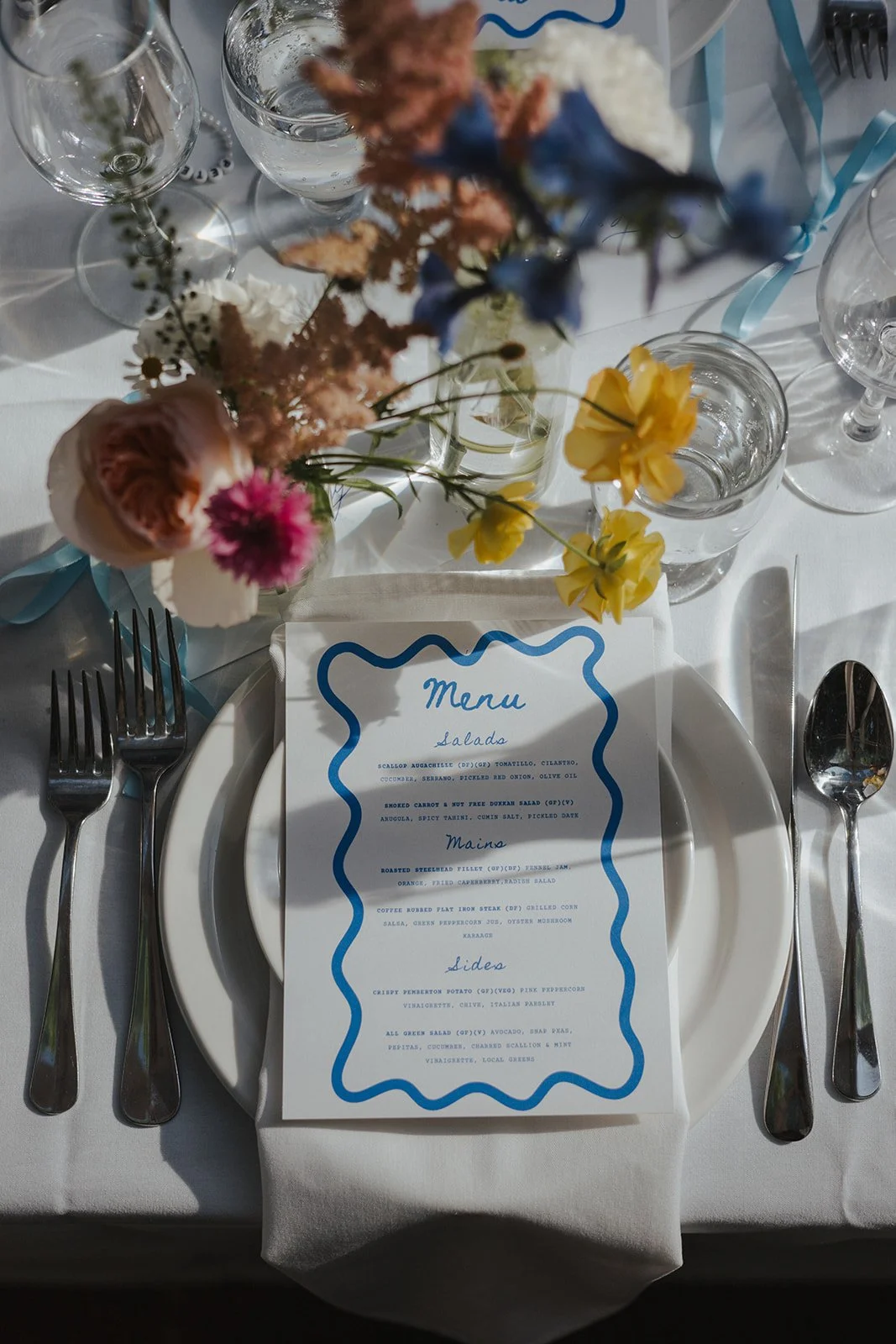 A formal table setting with a floral centerpiece and a printed menu, four forks to the left, and three knives and a spoon to the right, on a white tablecloth.