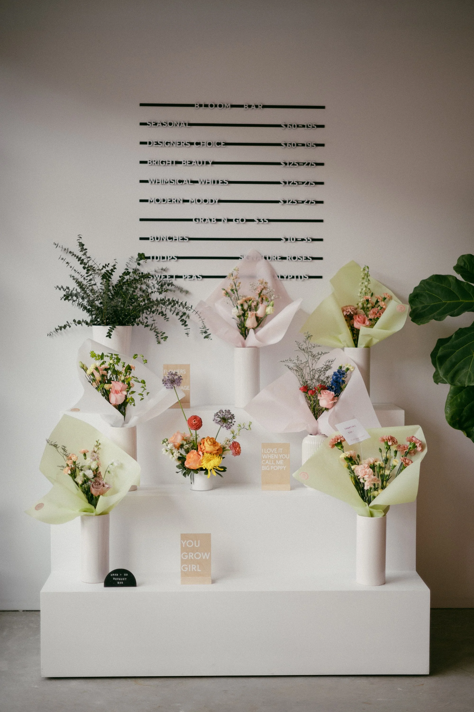 Display of various bouquets of flowers on a white stand in a floral shop, with a menu board on the wall above listing floral arrangements and prices.
