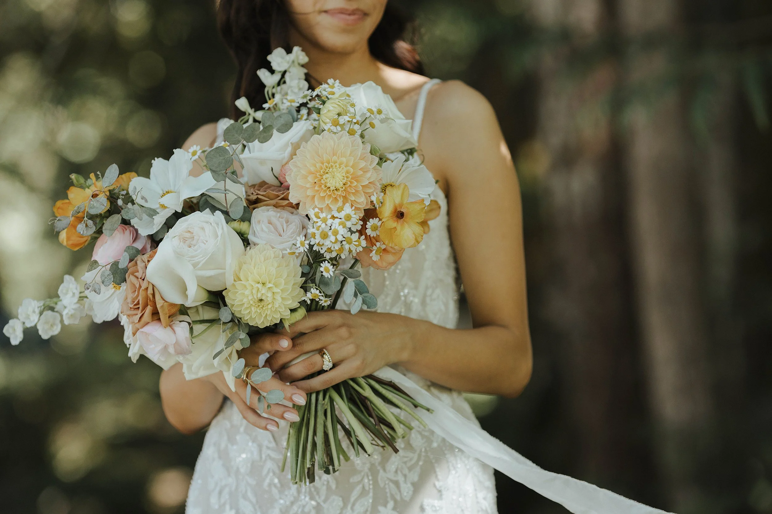 A woman in a wedding dress holding a bouquet of white and peach flowers, including roses, dahlias, and daisies, with greenery, outdoors with blurred trees in the background.
