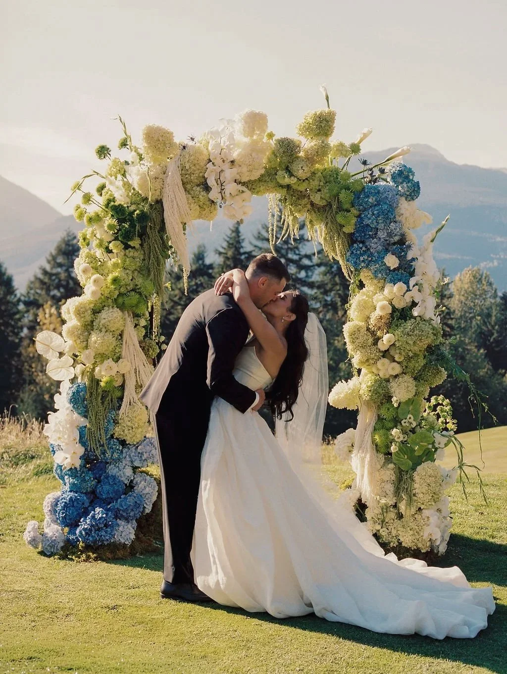 A bride and groom kissing under a large floral wedding arch outdoors.