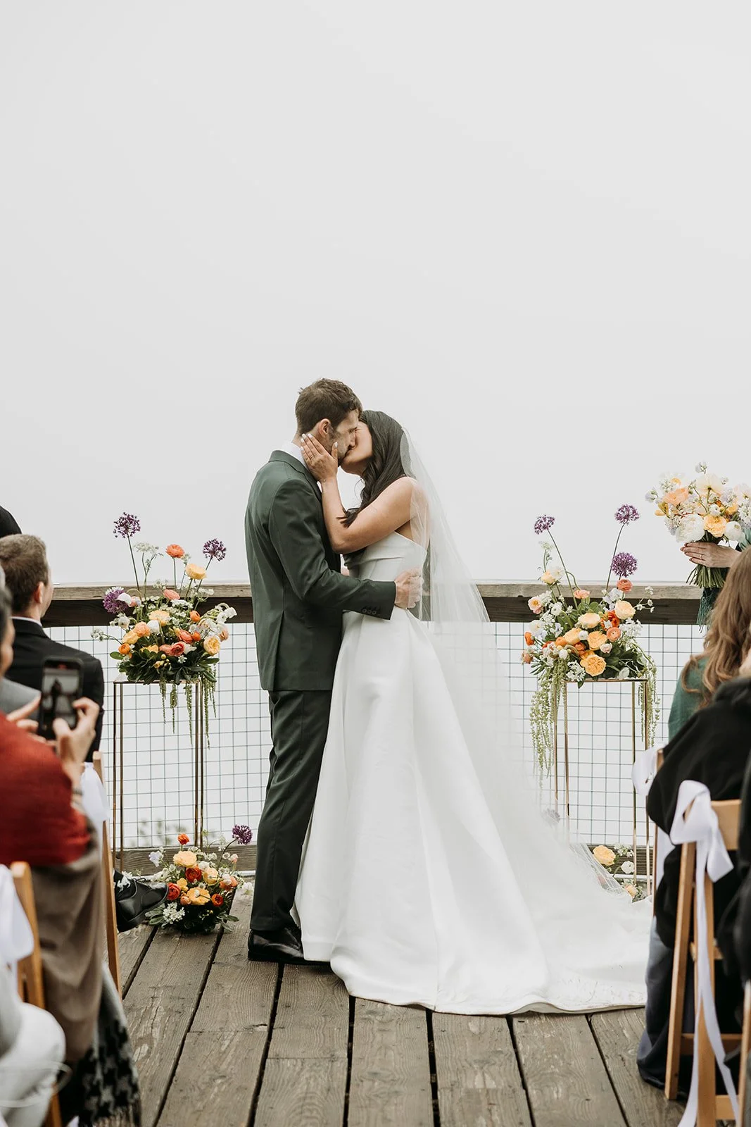 A bride and groom kiss during their wedding ceremony, standing in front of an officiant on a wooden platform decorated with colorful flowers.