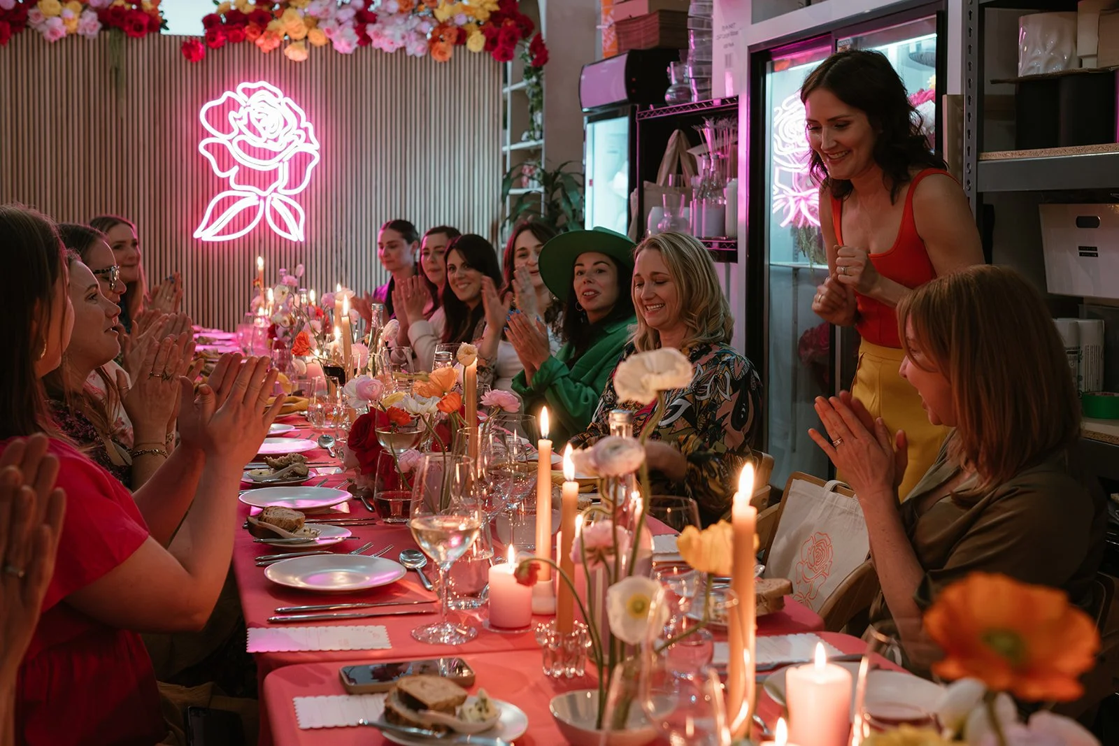 A celebration gathering with women seated around a long table decorated with pink flowers, candles, and food. One woman at the head of the table is standing and smiling, creating a joyful atmosphere. A pink neon rose sign is on the wall in the background.