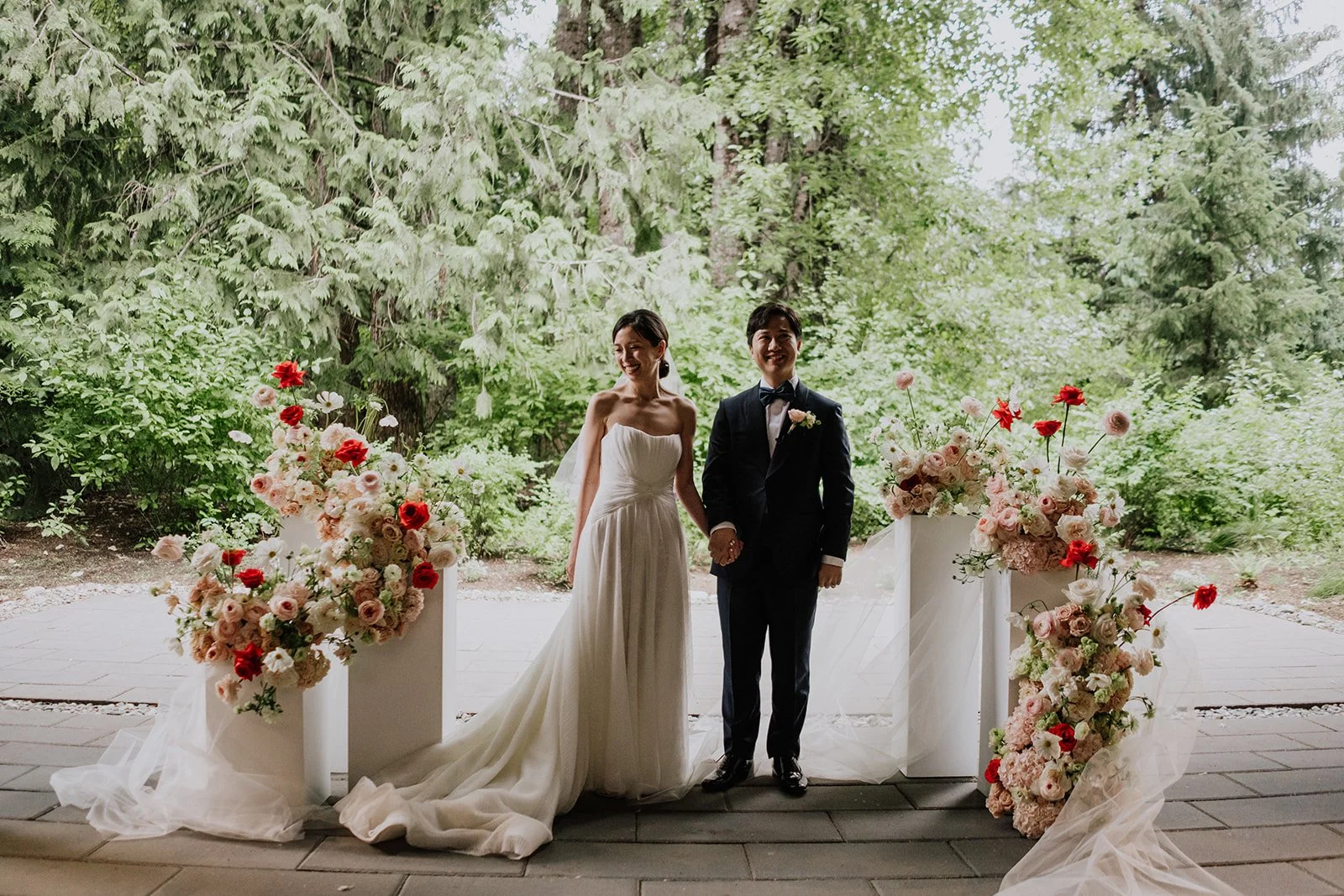 Bride and groom holding hands during wedding ceremony outdoors, surrounded by pink and red floral arrangements on pedestals, with lush green trees in background.