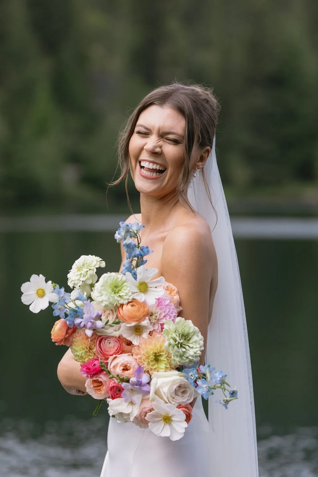 A joyful bride with a vivid smile holding a colorful bouquet of flowers outdoors near a body of water and green trees.