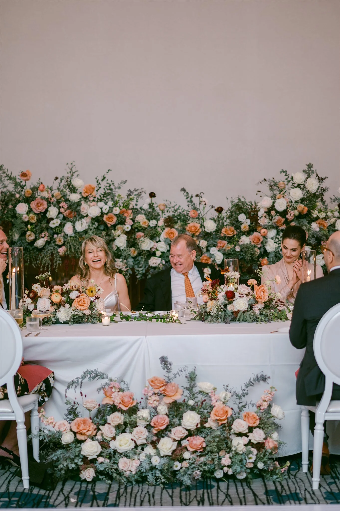 People sitting at a decorated wedding reception table, with flowers and candles, smiling and laughing, with a floral backdrop.