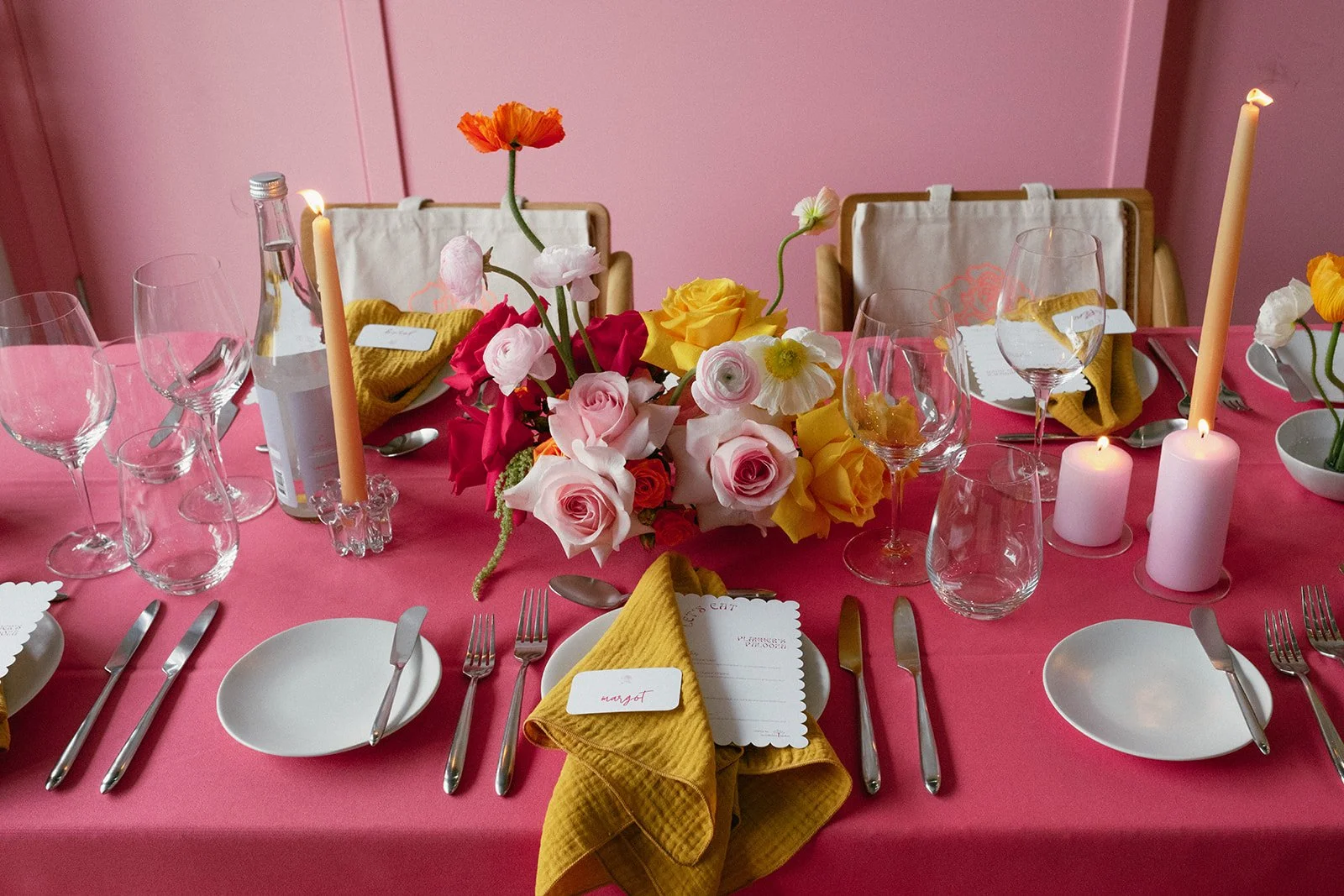 A pink tablecloth-covered table set for a dinner, featuring floral centerpieces with pink, yellow, white, and orange flowers, tall pink candles, and wine glasses. Gold utensils and yellow cloth napkins with name tags are also arranged on the table.