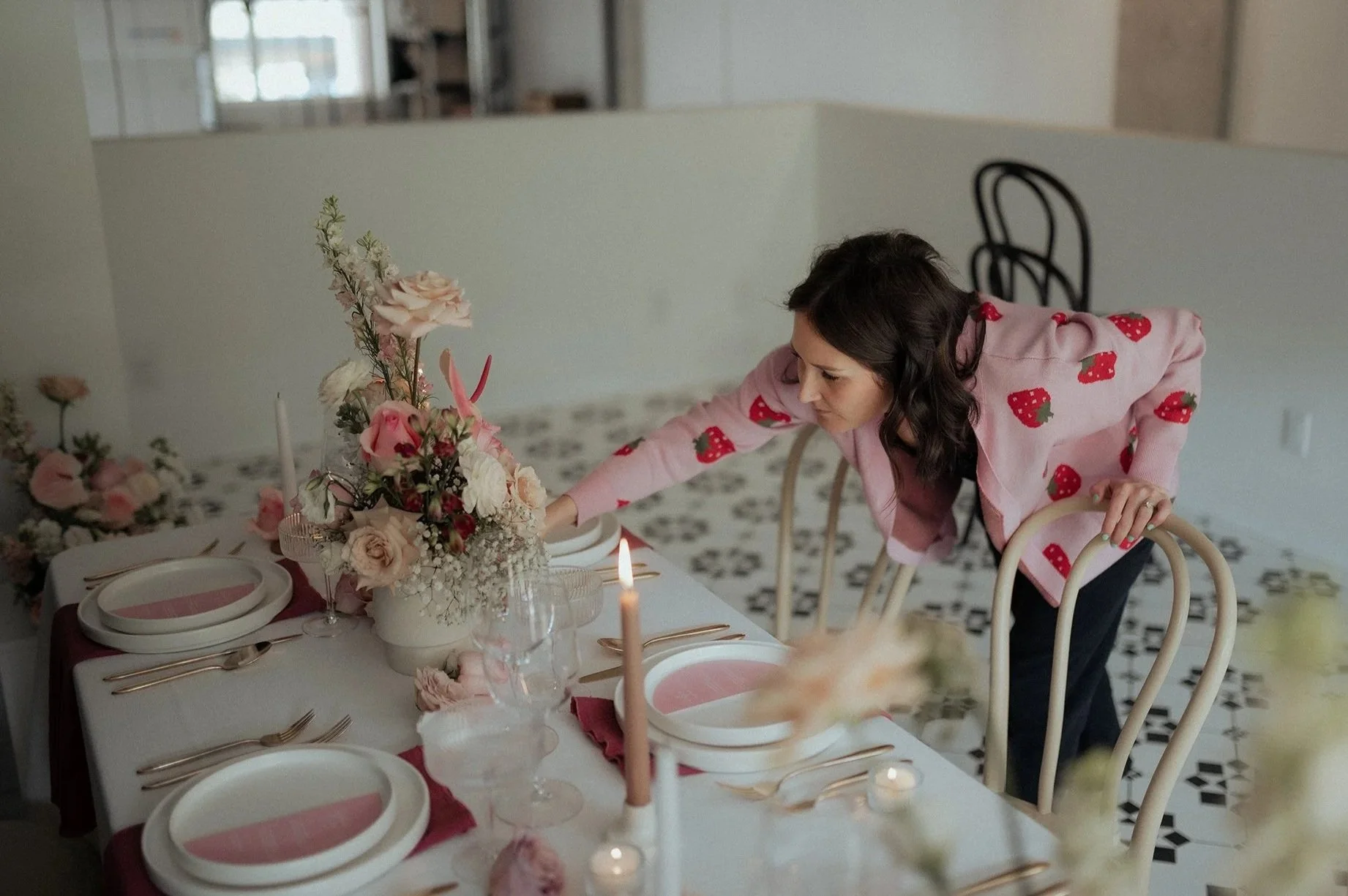 Woman arranging a floral centerpiece on a decorated dining table with plates, cutlery, candles, and flowers in a bright room.