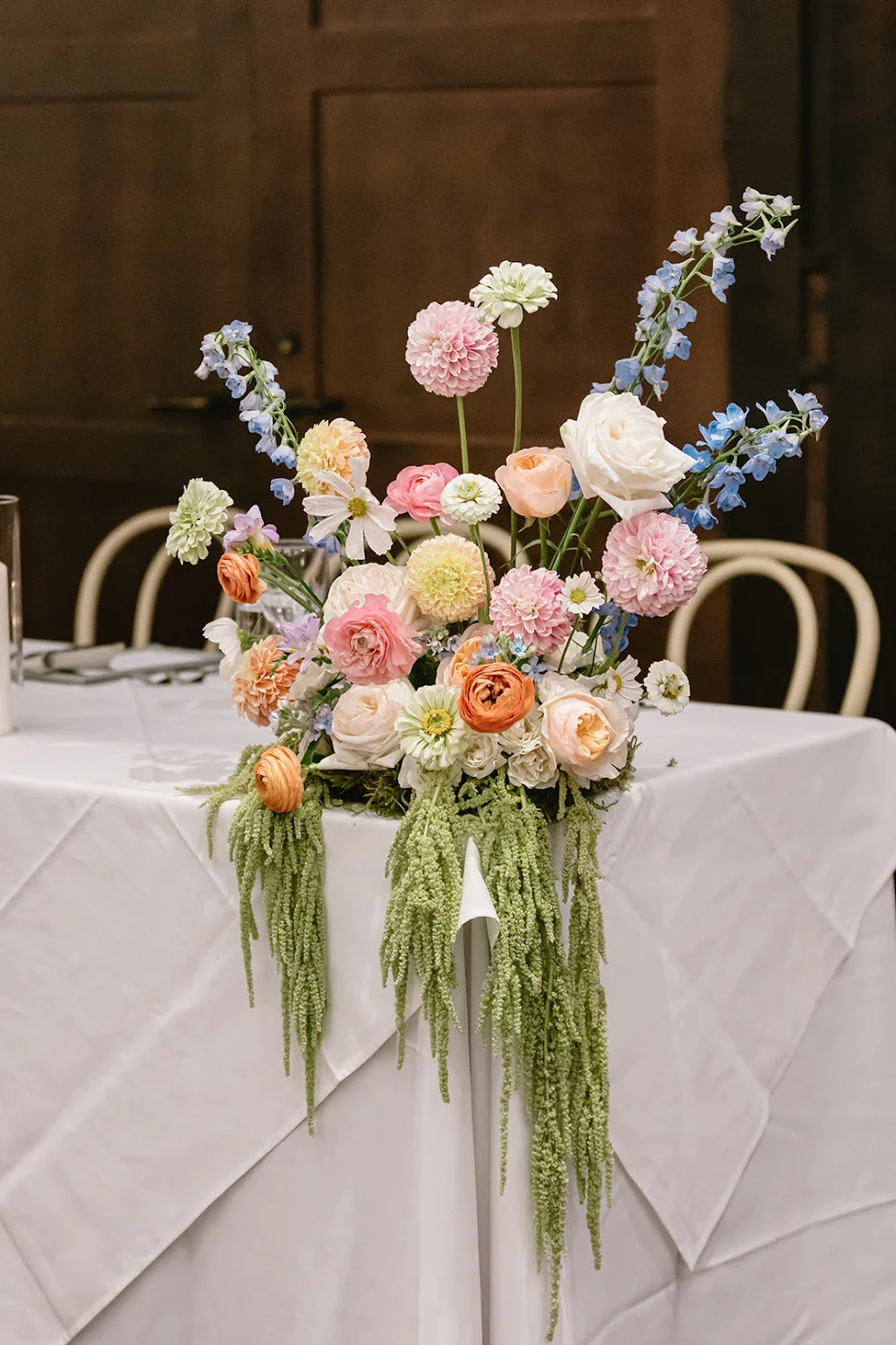 A floral centerpiece with pink, white, and peach flowers, including roses, dahlias, and delphiniums, arranged on a table with a white tablecloth.