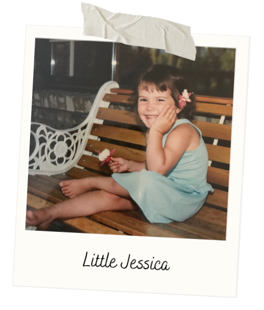 A young girl with short brown hair, wearing a light blue dress, sitting on a wooden bench outdoors, holding a small flower, smiling with her hand on her cheek, with a white ornate bench behind her. The photo is labeled "Little Jessica" at the bottom.