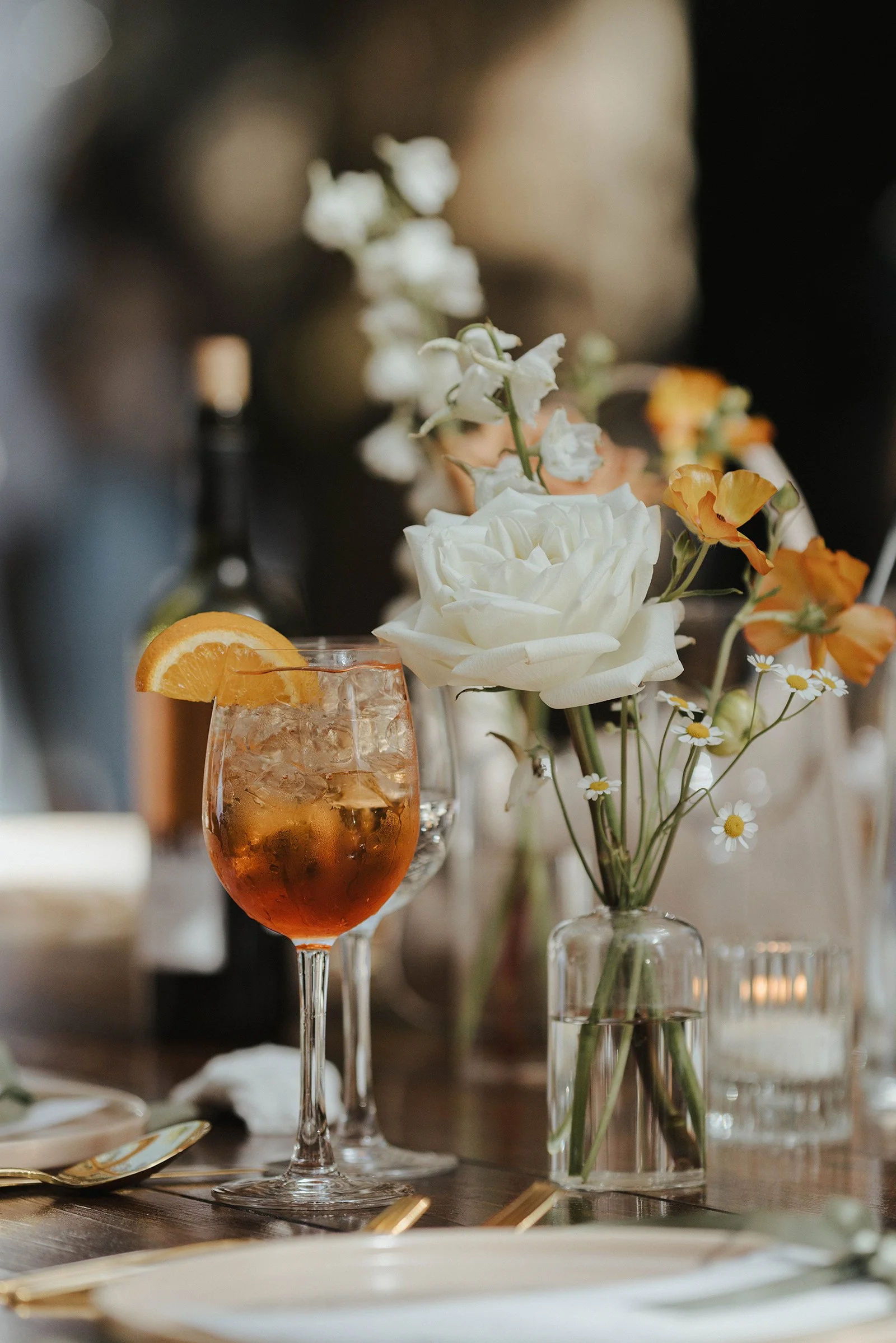 A table centerpiece with white, orange, and small white daisies in a clear glass vase, beside a tall glass of iced orange beverage with an orange wedge, and surrounded by candles, plates, and cutlery.