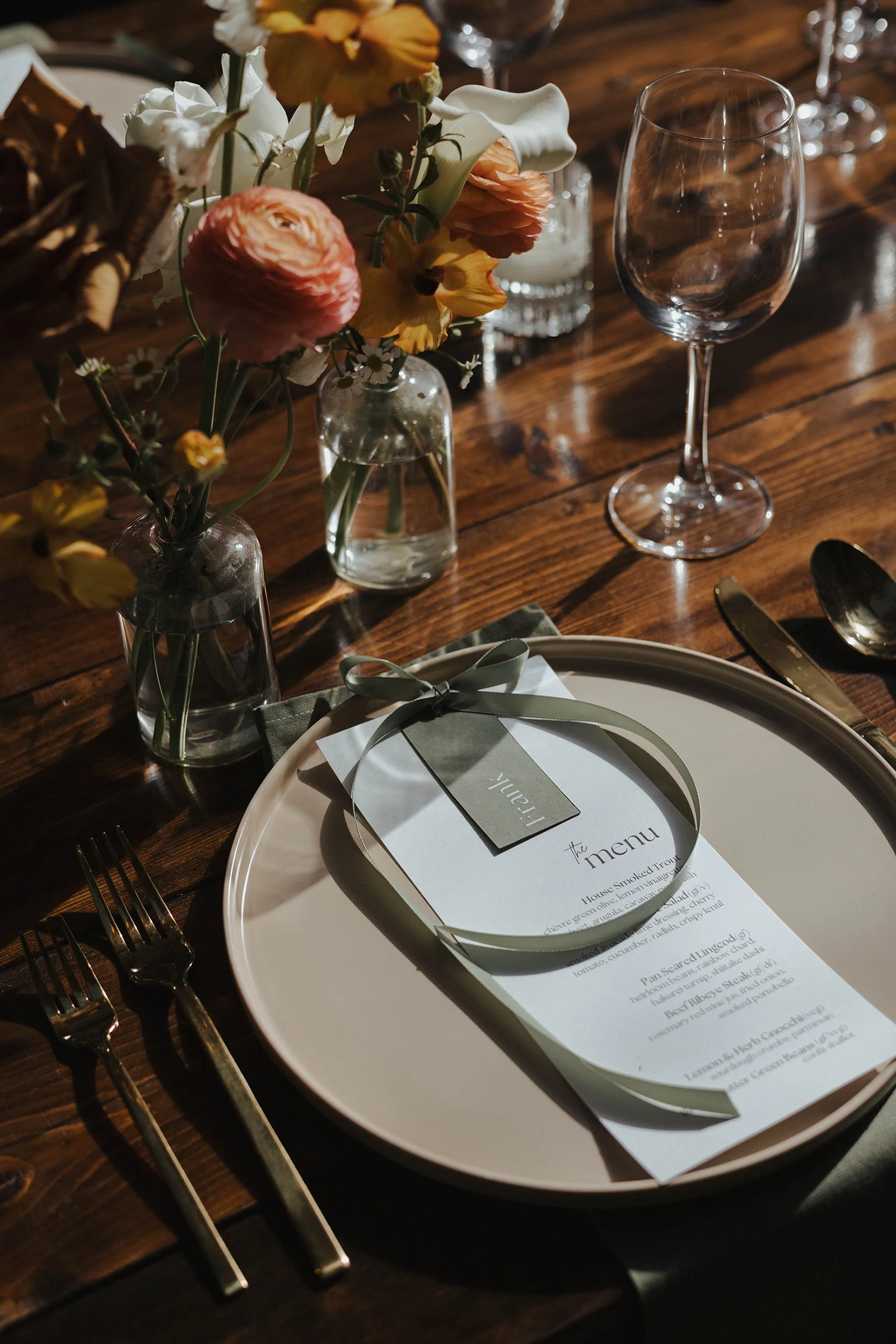 Elegant table setting with a menu on a white plate, surrounded by silverware, flower vases with pink and yellow flowers, and empty wine glasses at a dinner event.