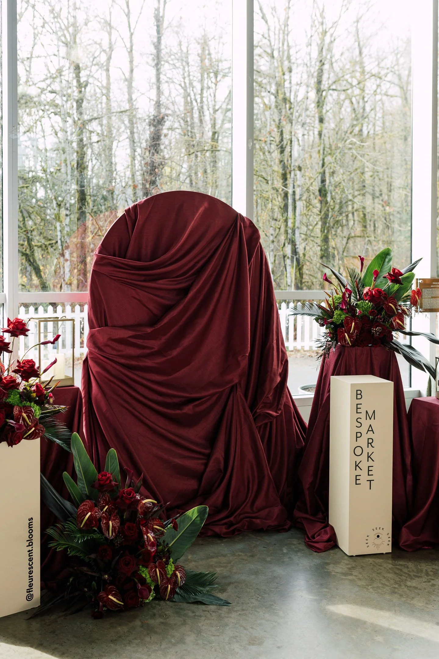 A burgundy velvet-covered display with a curved top, surrounded by burgundy floral arrangements, inside a room with large windows showing leafless trees outside. There is a white rectangular sign with black text reading 'BESPOKE MARKET' and a small logo at the bottom.