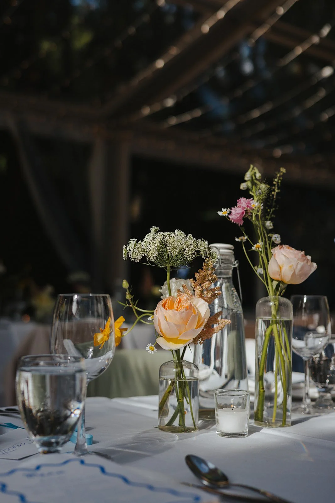 Table with floral centerpieces in glass vases, wine glasses, water glasses, a silver spoon, and small white candles in glass holders at an outdoor event.