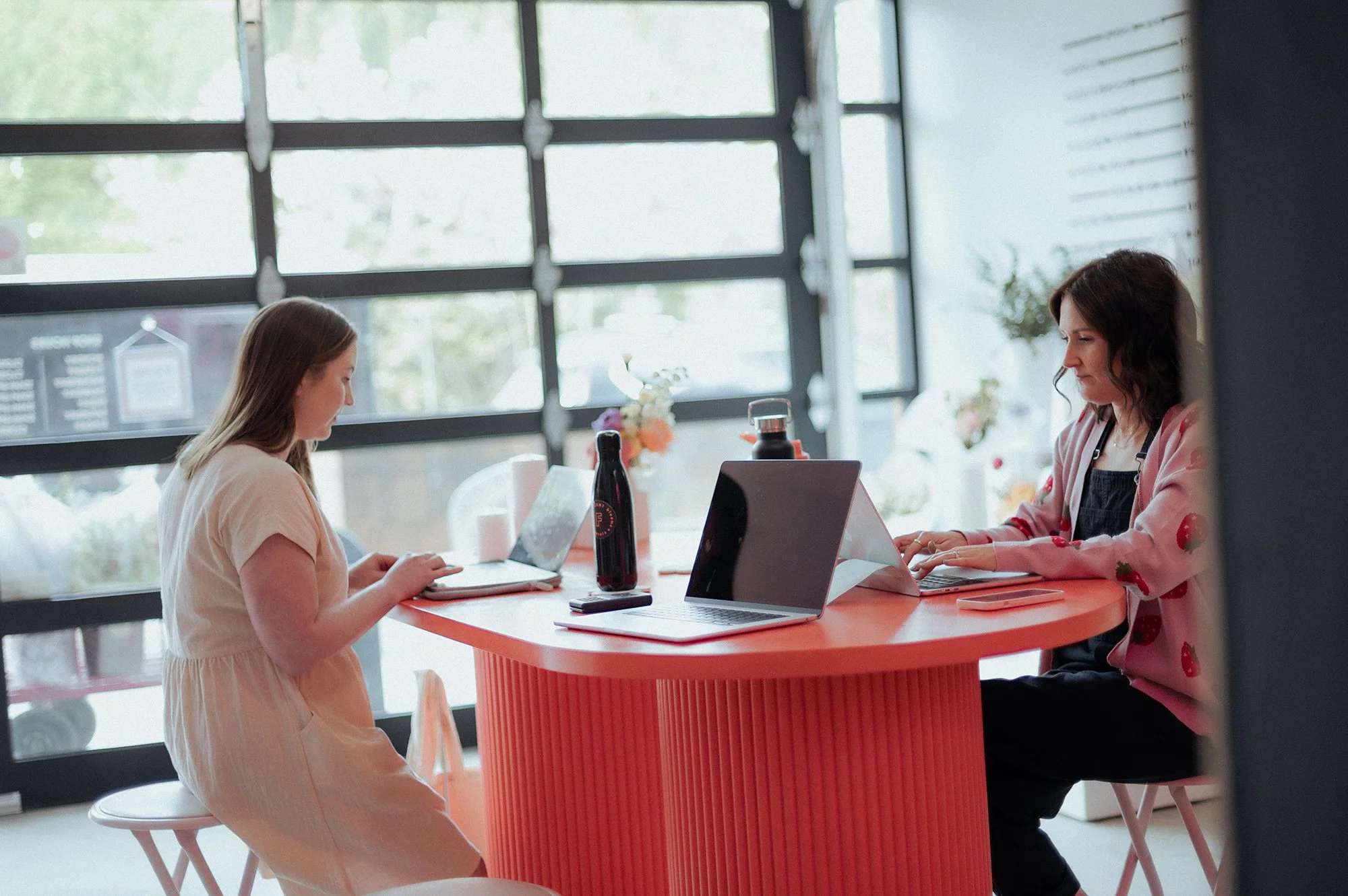 Two women sitting at an orange table in a bright cafe, working on laptops and tablets.