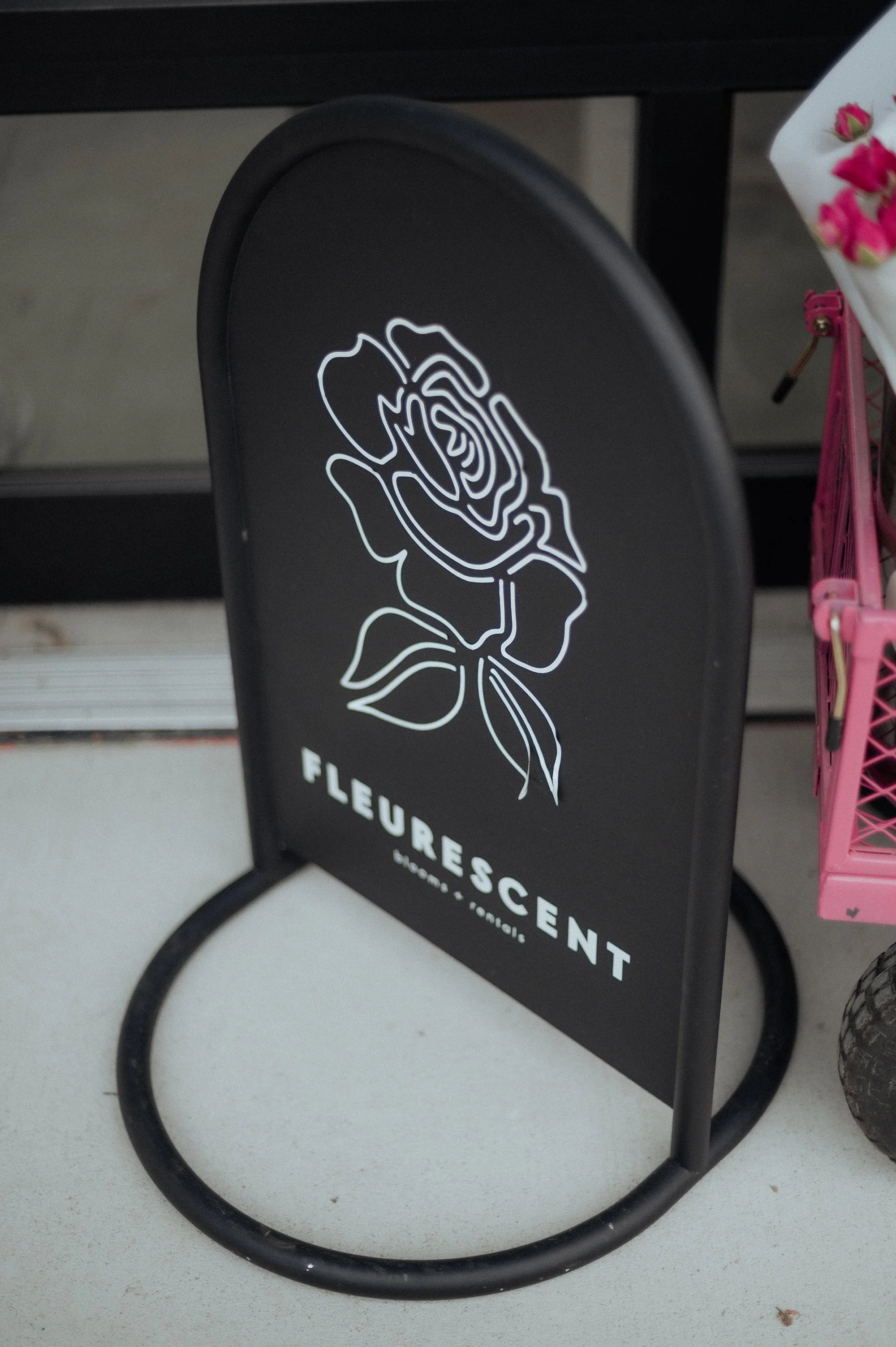 Black signboard with a stylized white outline of a rose and the text "FLEURESENT, HOME & LOVE" beneath the rose.
