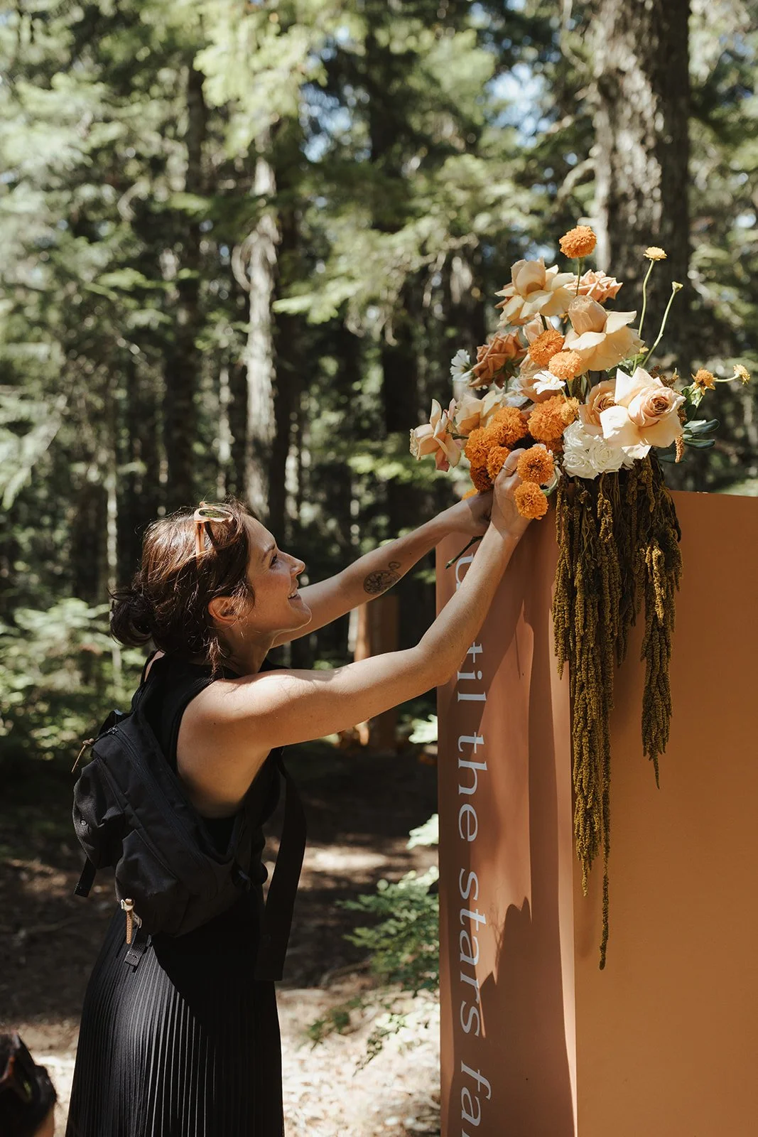 A woman with sunglasses on her head and a black backpack adjusts a floral arrangement on a peach-colored stand in a forest setting.
