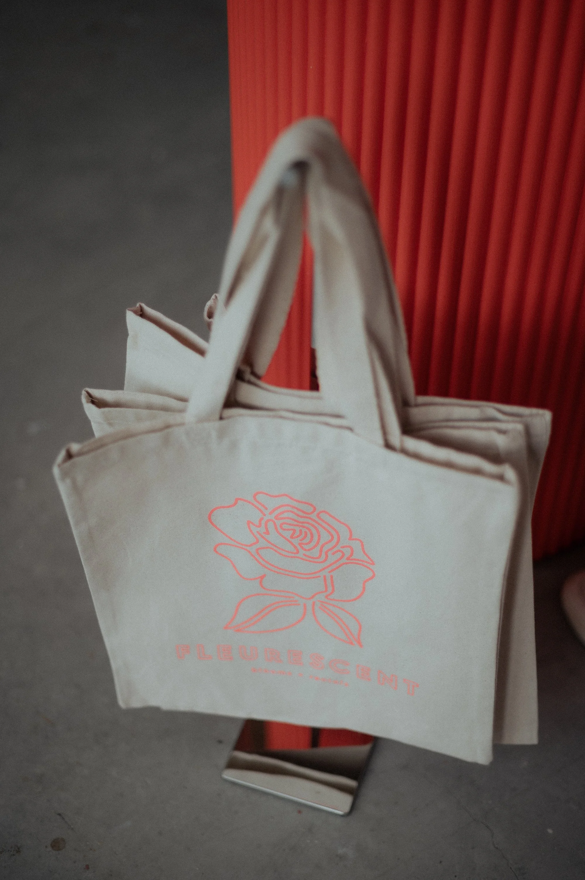 A beige tote bag with a rose design and the word 'FLEURS CENT' printed on it, hanging on a mirror display stand in front of a red textured background.