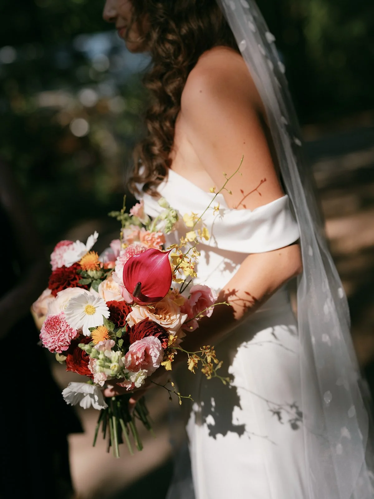 A bride holding a colorful bouquet of flowers outdoors, with sunlight casting shadows on her white dress and veil.