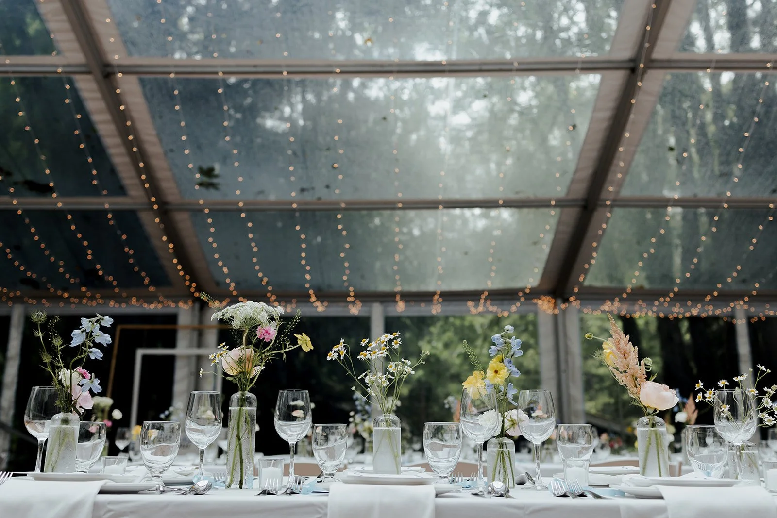 Elegant banquet table set under a glass ceiling with string lights, decorated with flower arrangements in vases, wine glasses, and tableware for a celebration or wedding reception.