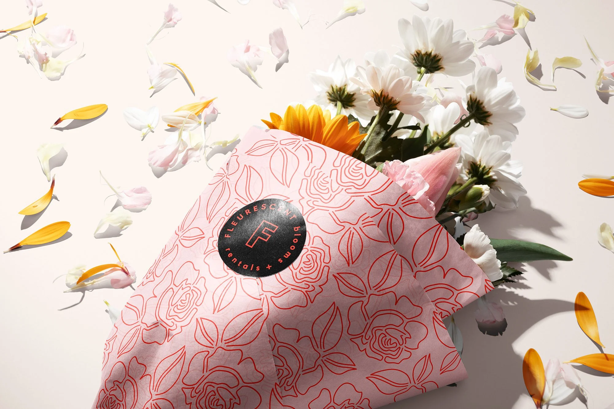 A bouquet of white, pink, and yellow flowers wrapped in pink floral-patterned paper with a black sticker labeled 'FLEURESSENCE' on a white background with scattered flower petals.