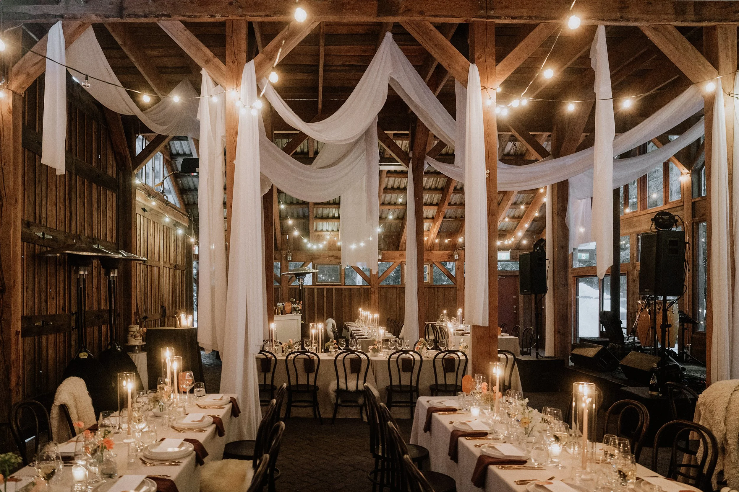 An elegantly decorated rustic event space with wooden walls and ceiling, draped white fabric, and string lights, set up for a wedding reception or special occasion with long tables, candles, flowers, and seating.