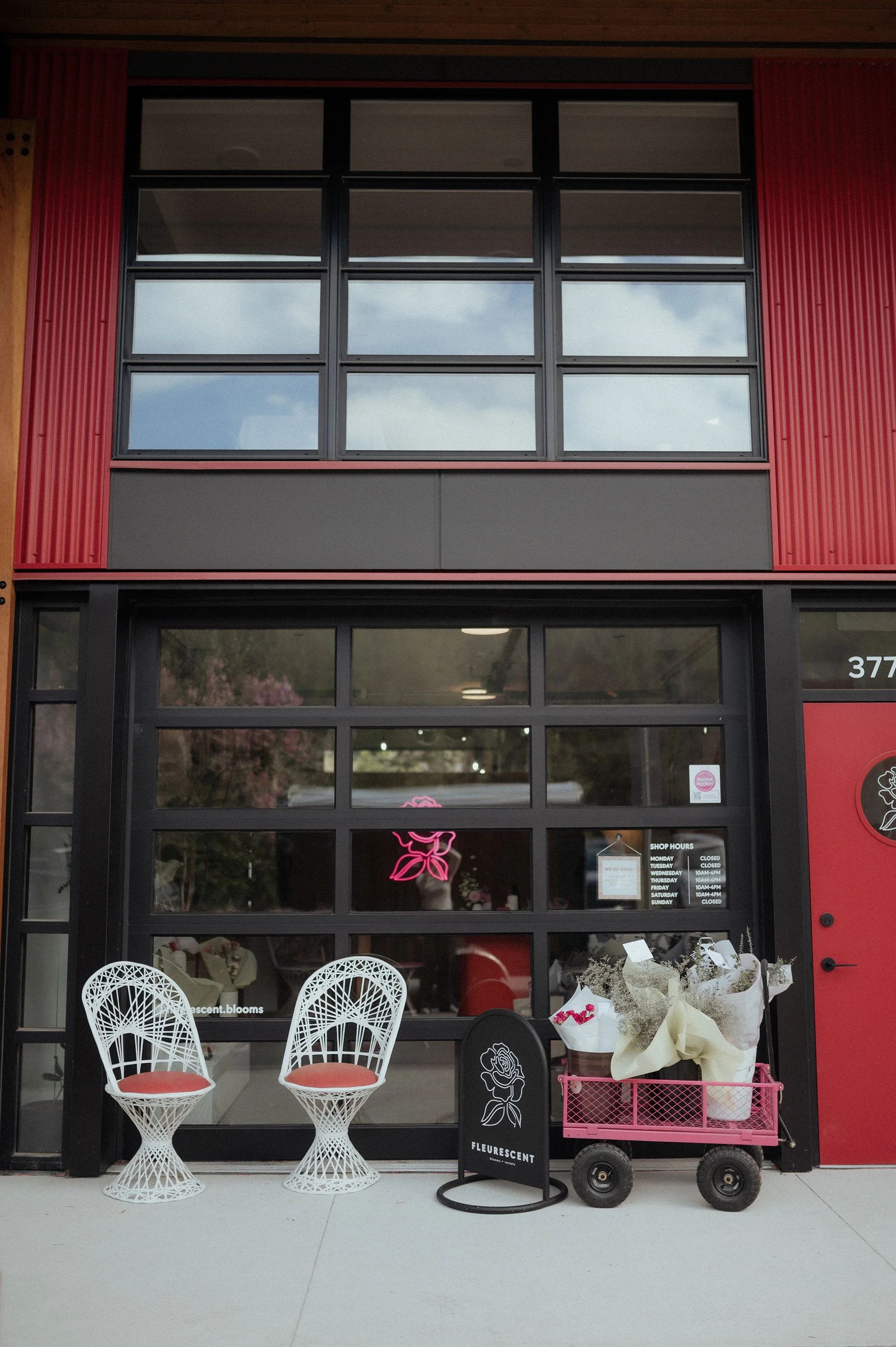 Flower shop storefront with two white wire chairs with red cushions outside, pink floral arrangements in a cart, and neon pink flower sign in the window.