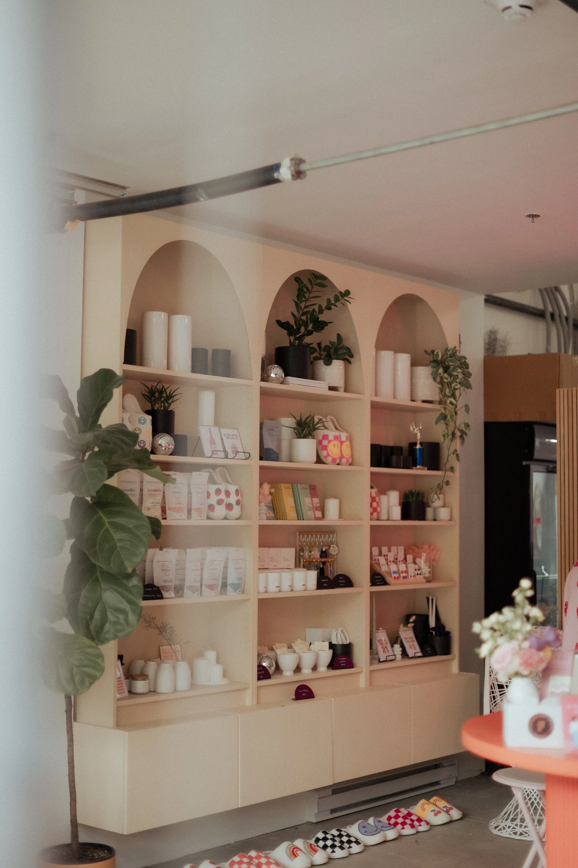 Interior of a store with shelves displaying potted plants, candles, and decorative items, part of a round orange table with flowers, and patterned slippers on the floor.