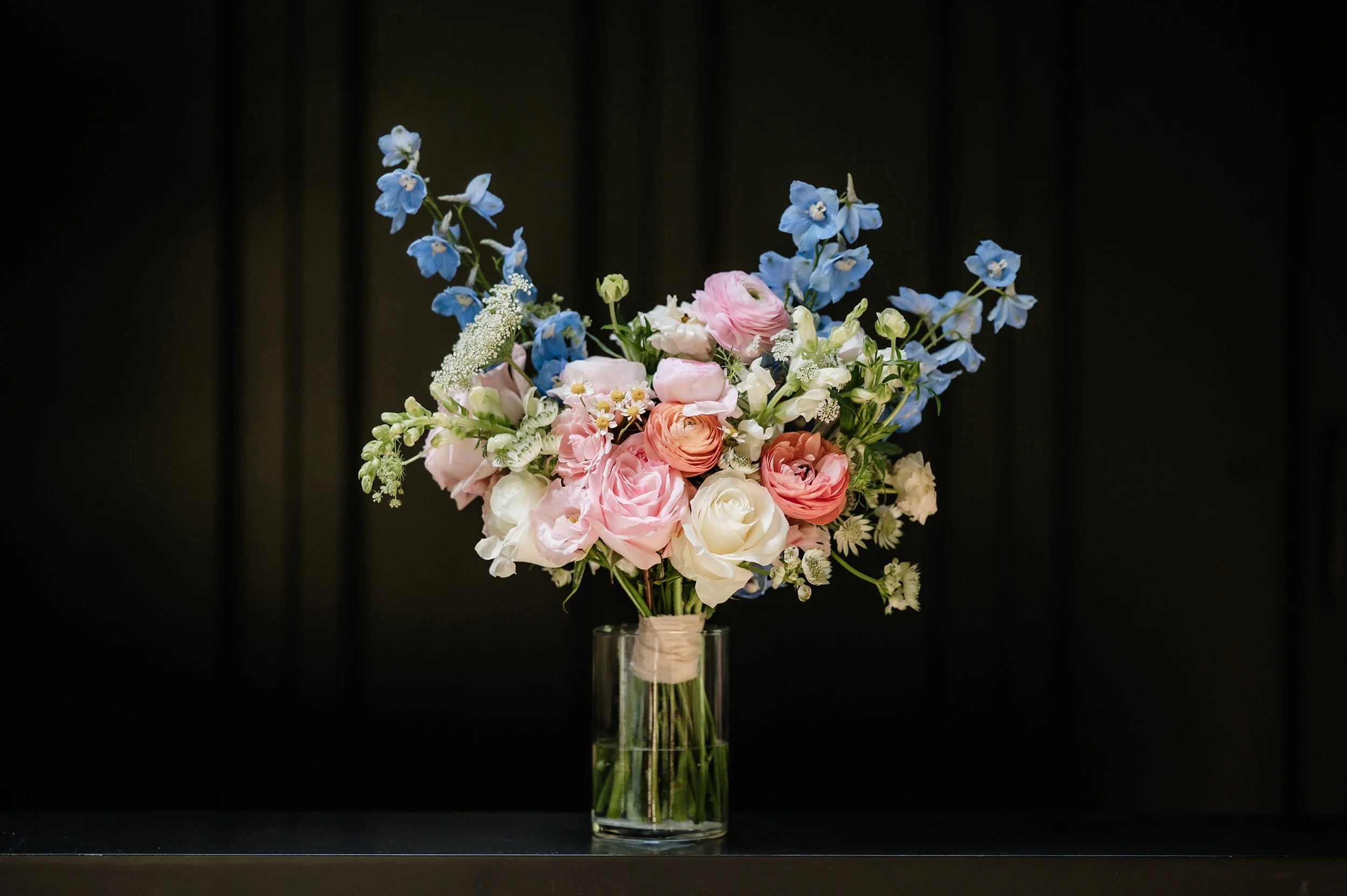 A glass vase with a bouquet of pink, white, and peach roses, blue delphiniums, and small white filler flowers, set against a dark background.
