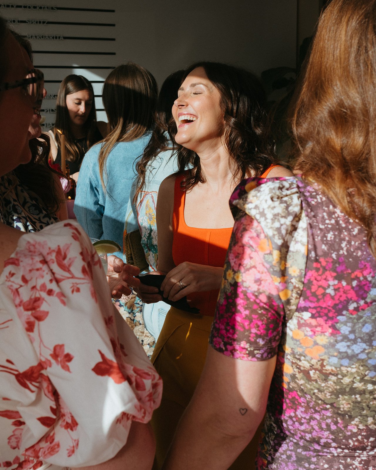Group of women laughing and talking at a social gathering, some holding drinks, in a well-lit indoor setting.