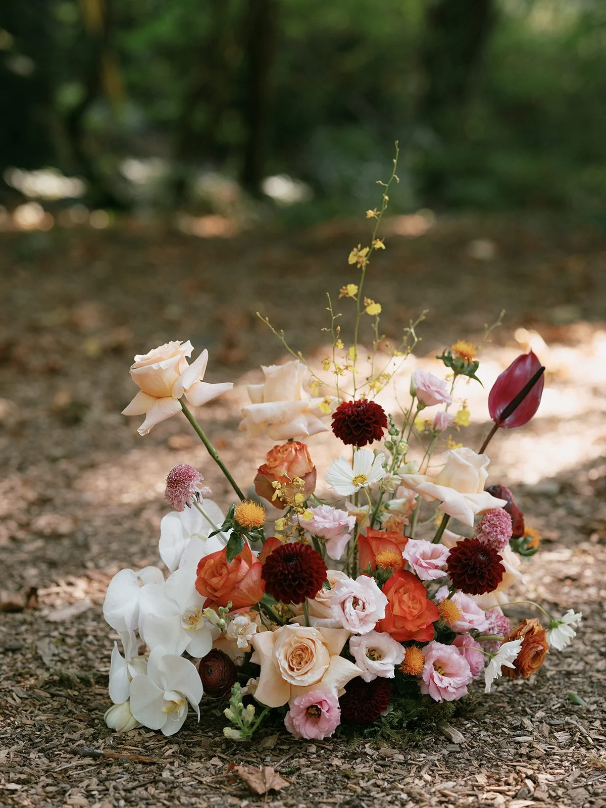 A colorful bouquet of various flowers including roses, dahlias, calla lilies, anthuriums, and other blooms, placed on a wooded ground with blurred background.