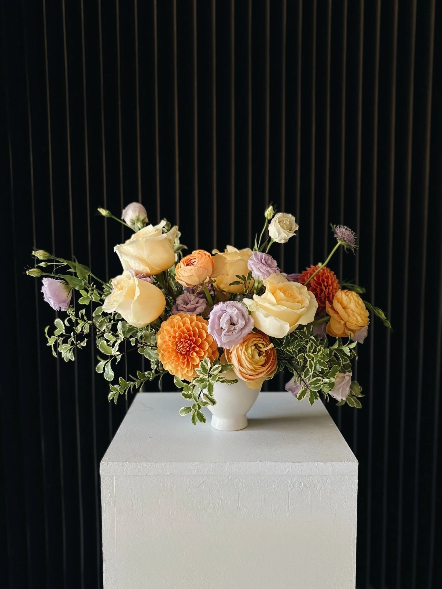 A floral arrangement with cream, orange, and light purple flowers in a white vase on a white pedestal against a black striped background.