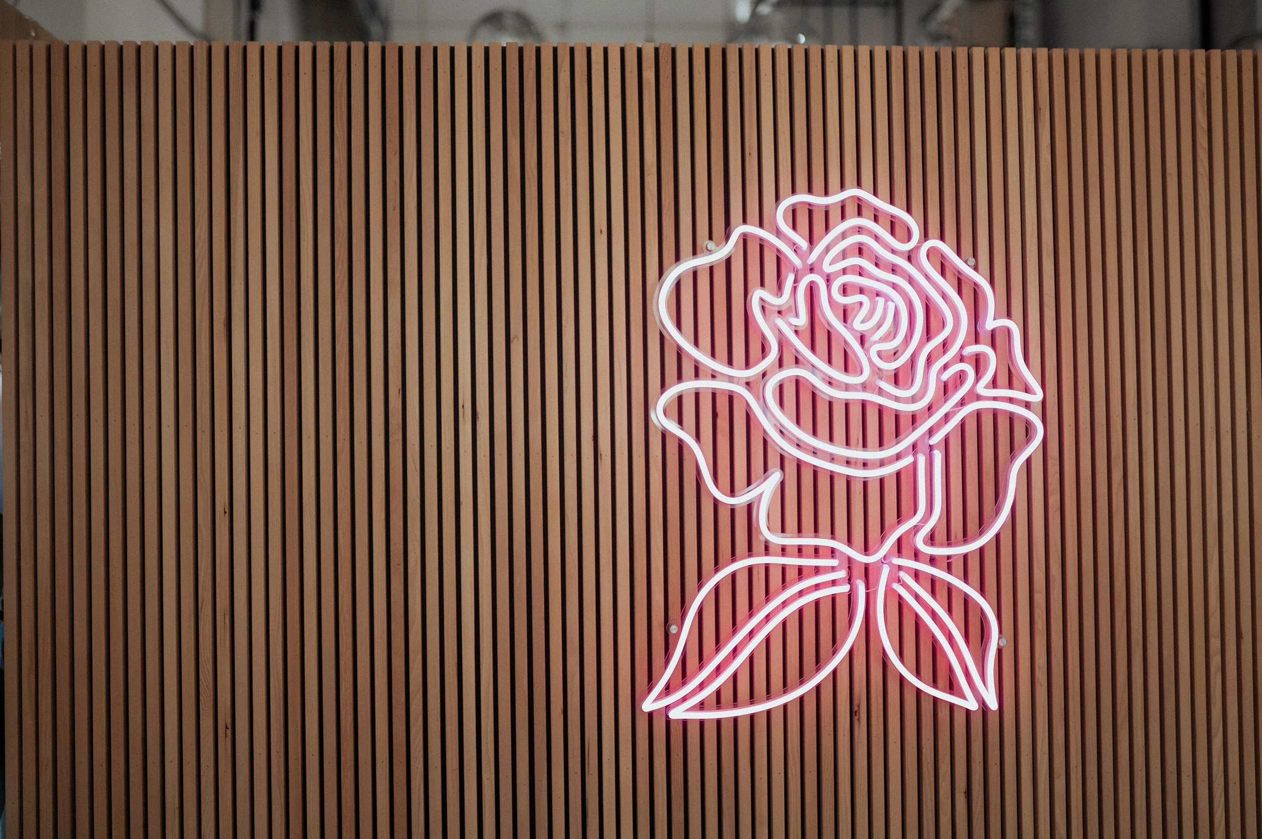 Pink neon outline of a rose flower mounted on a wooden slat wall.