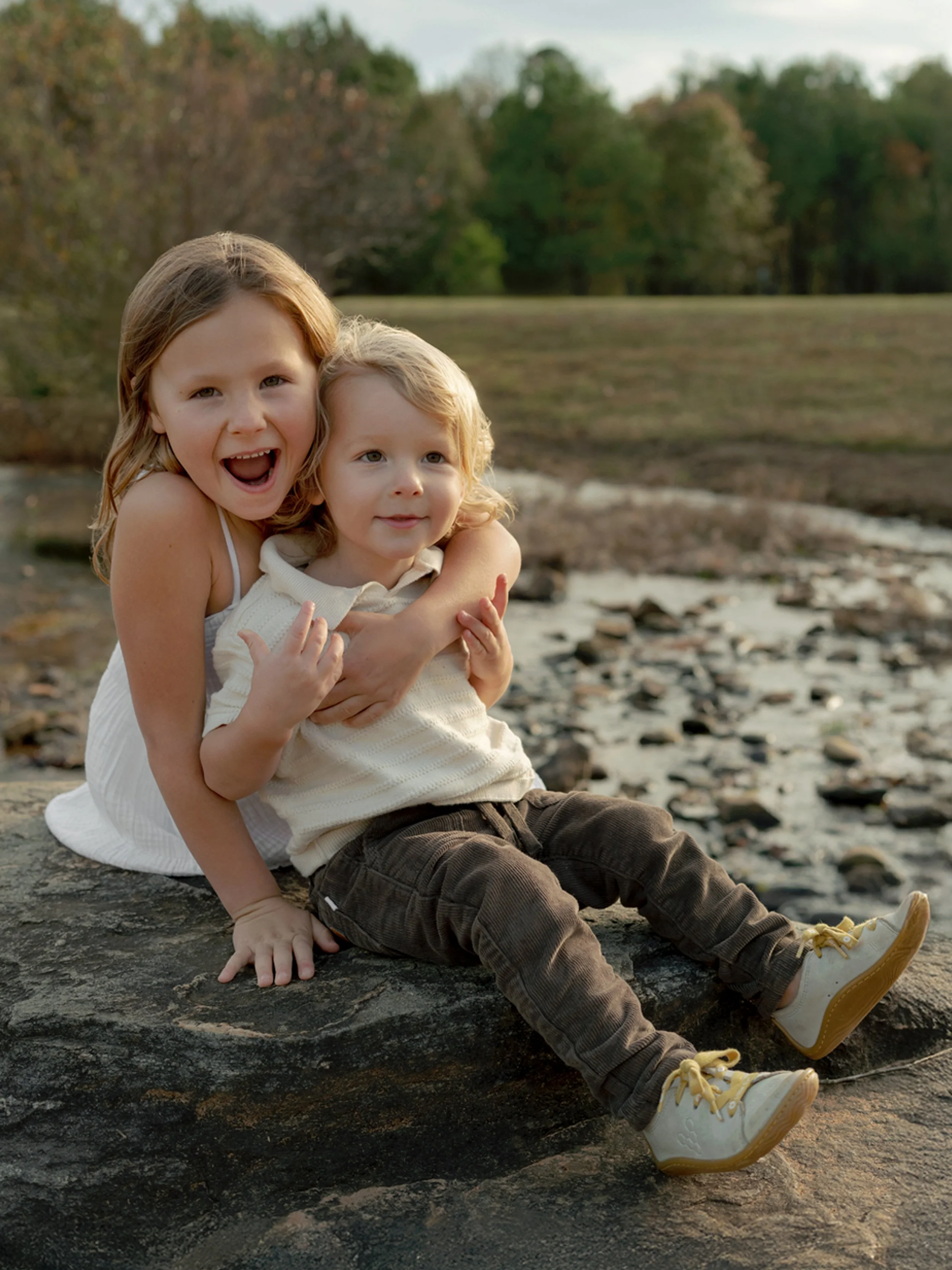 Two young children, a girl and a boy, sitting on a rock outdoors near a river with trees in the background. The girl is smiling widely with her mouth open, hugging the boy who is looking off to the side.
