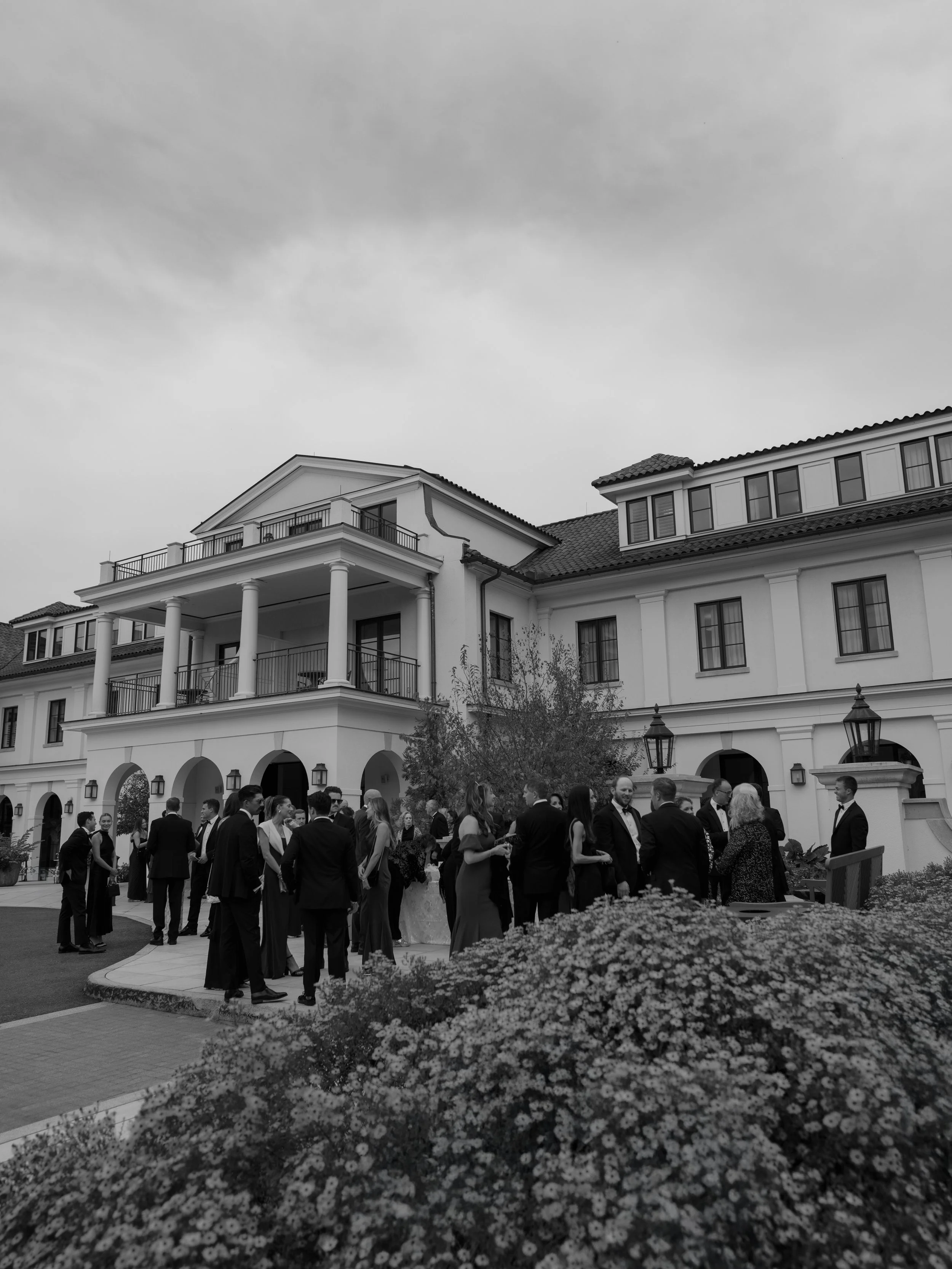 Group of people in formal attire gathered outside a large elegant building with balconies, during daytime, for a social event or wedding.