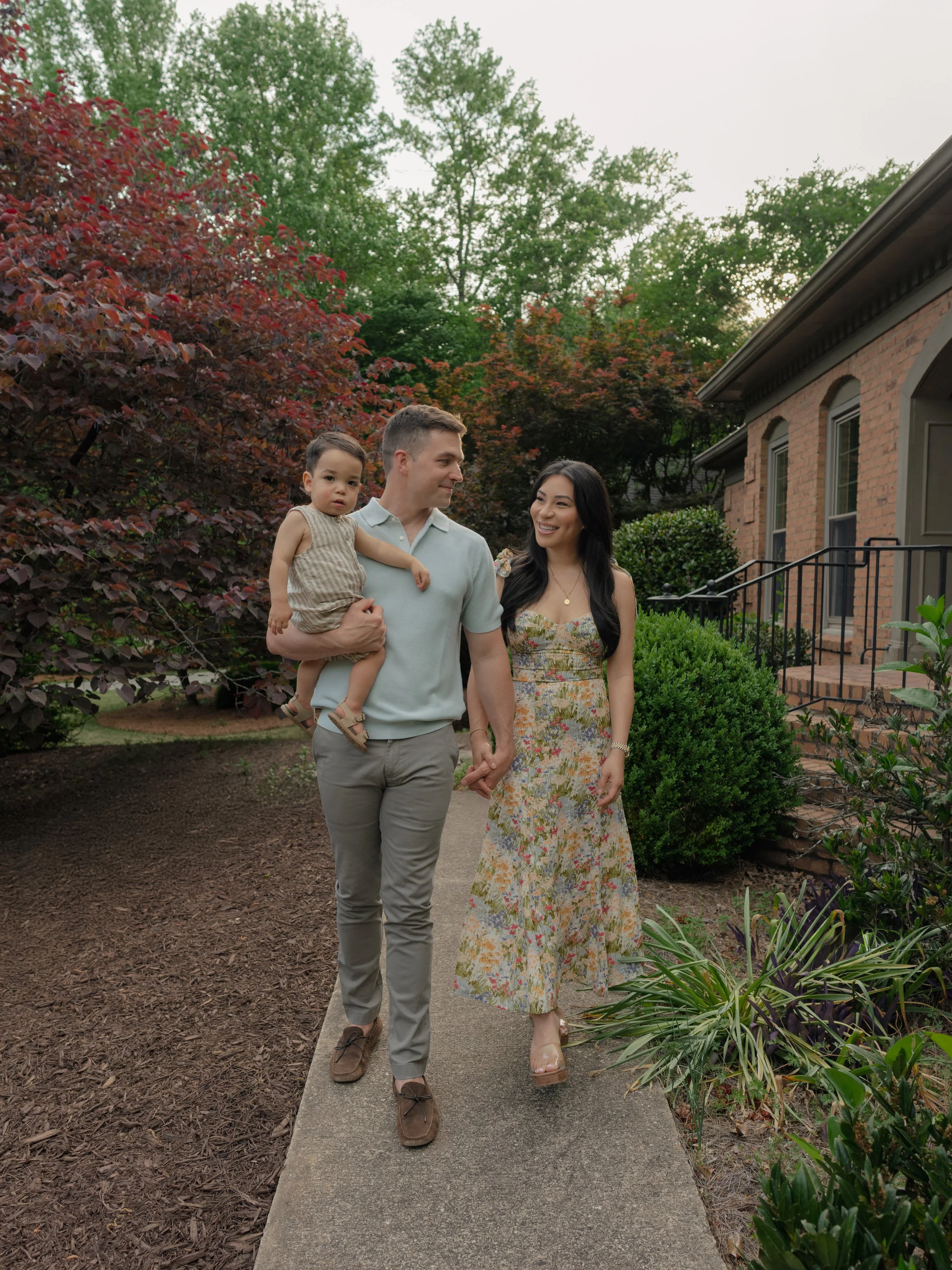 A couple walking in a garden with a young child. The man is carrying the child who is looking at the camera. The woman is smiling and holding the man's hand. The garden has green and red foliage with a brick house in the background.