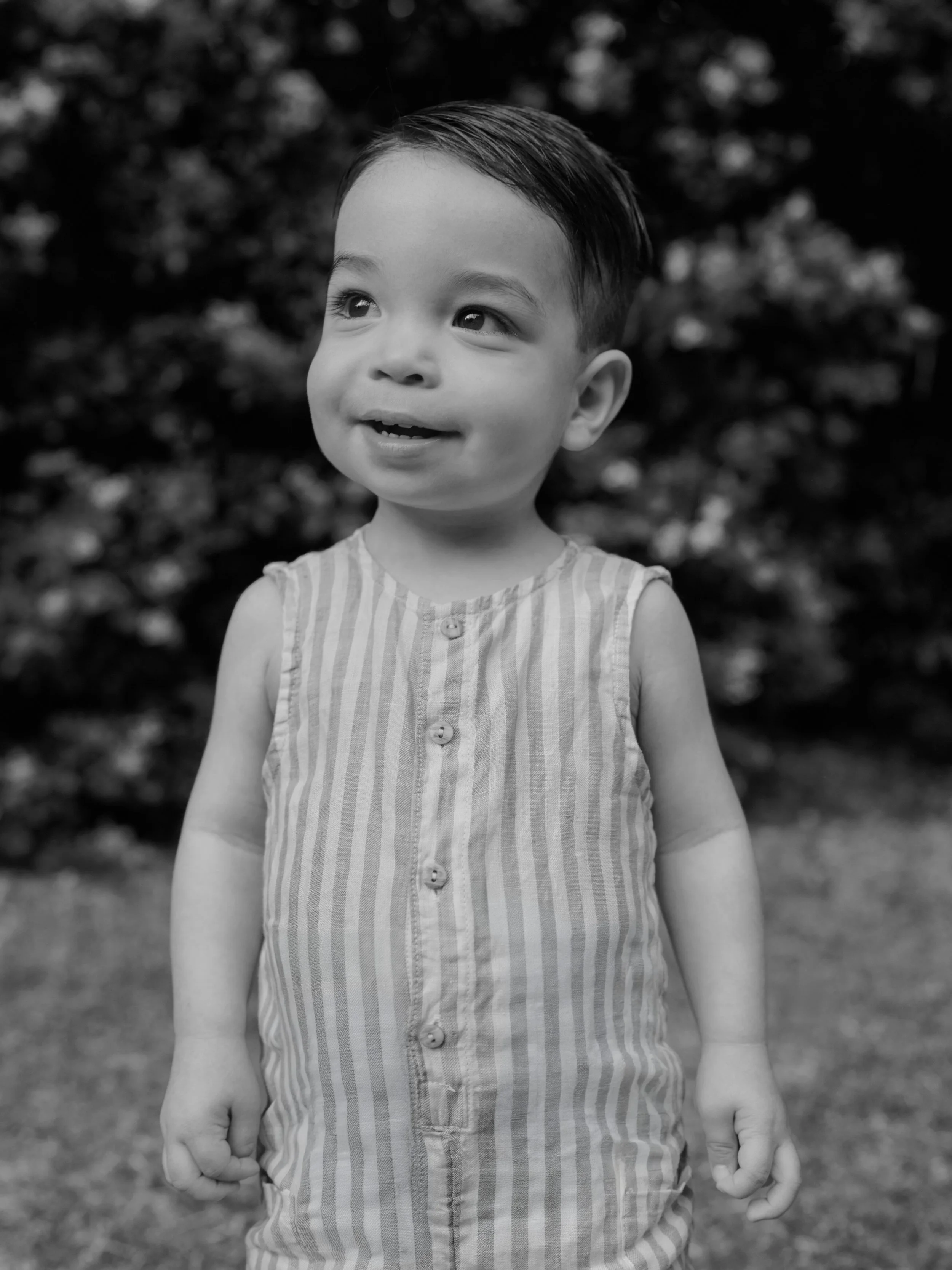 A young boy with short, slicked-back hair wearing a sleeveless, button-up shirt standing outdoors with blurred foliage in the background.