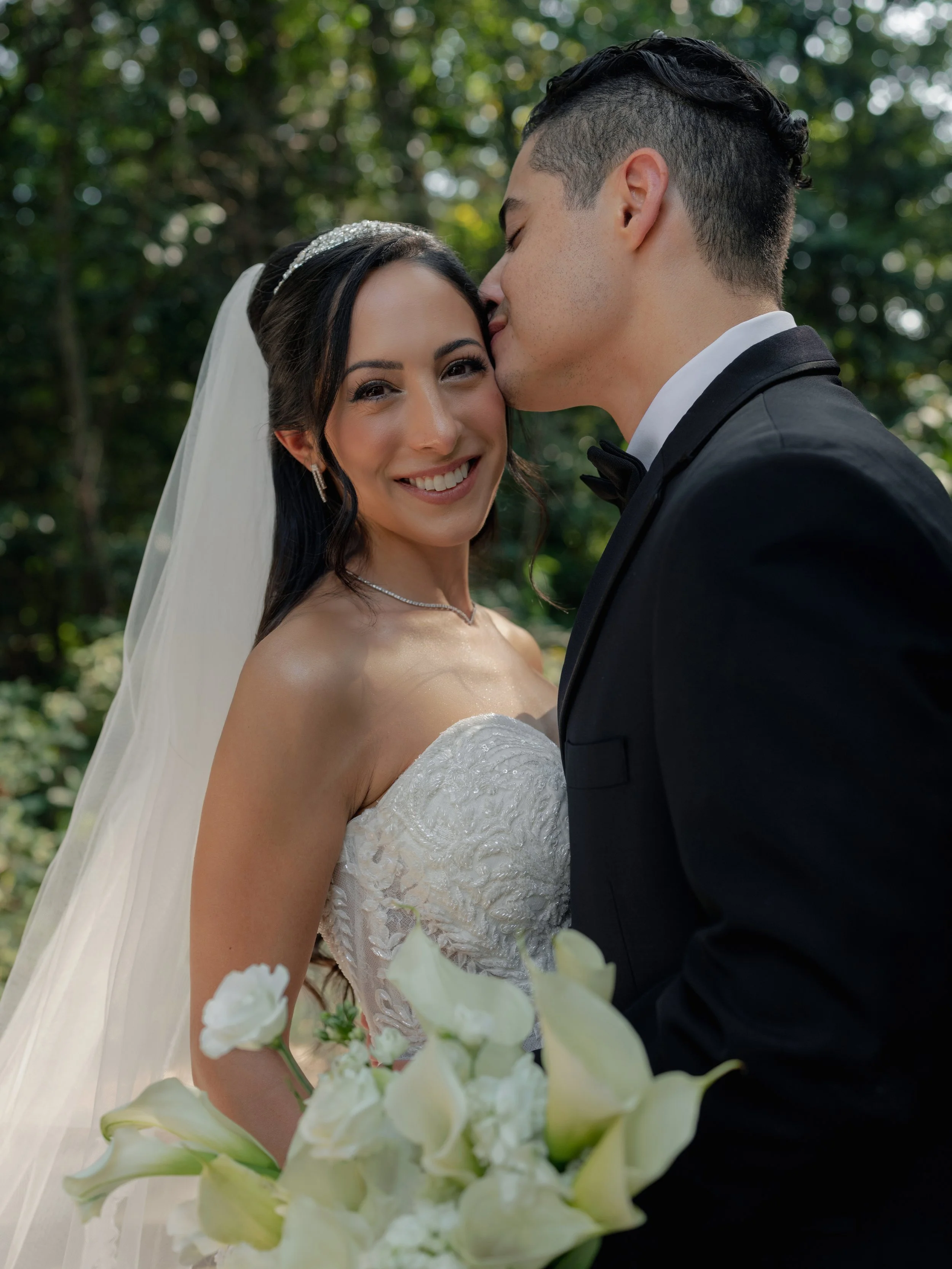 A bride and groom on their wedding day, with the groom kissing the bride's forehead and the bride smiling at the camera, holding a bouquet of white flowers outdoors.