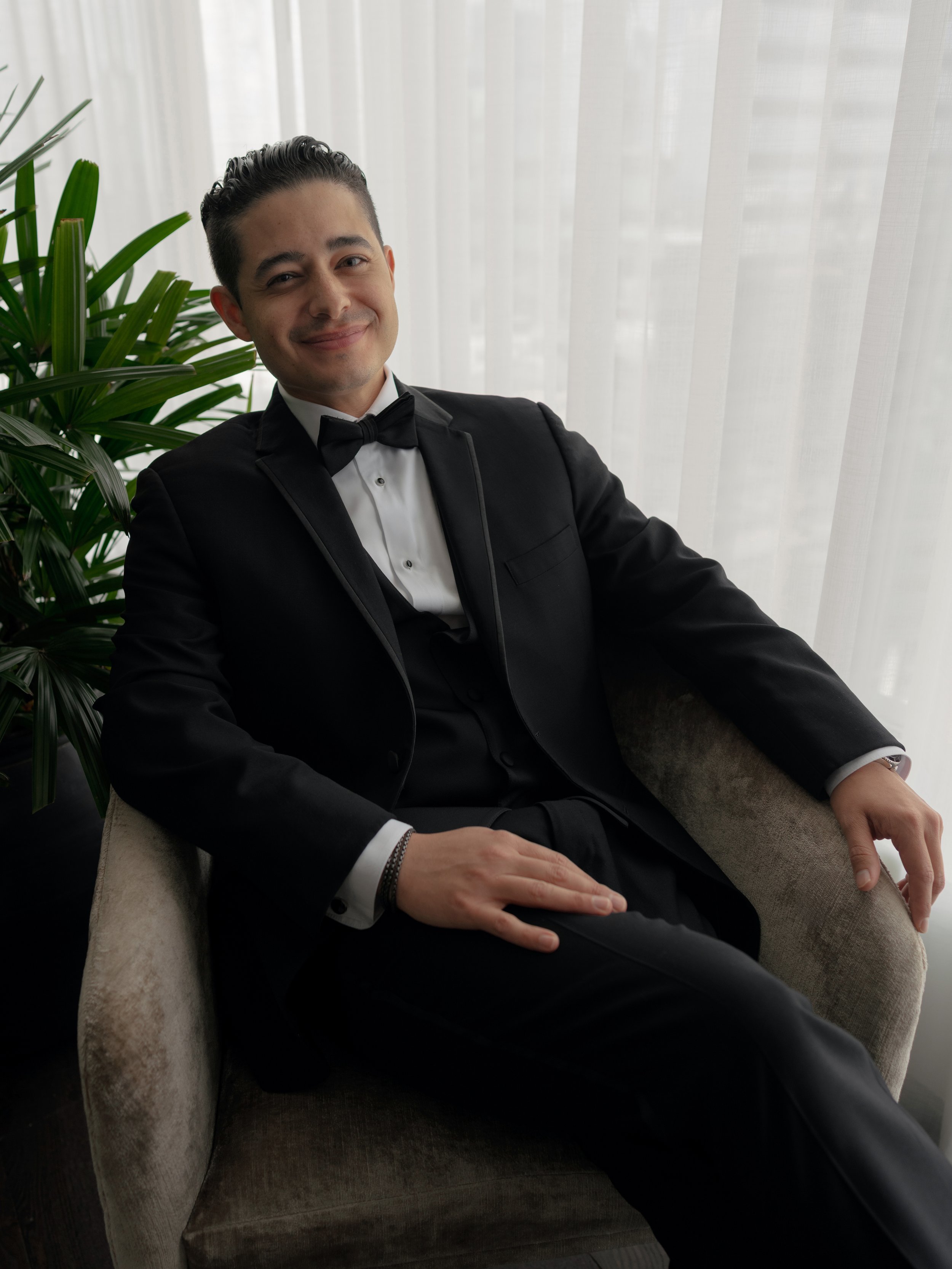 A man in a black tuxedo with a bow tie, sitting in a beige armchair, smiling, with a large green plant and white curtains in the background.