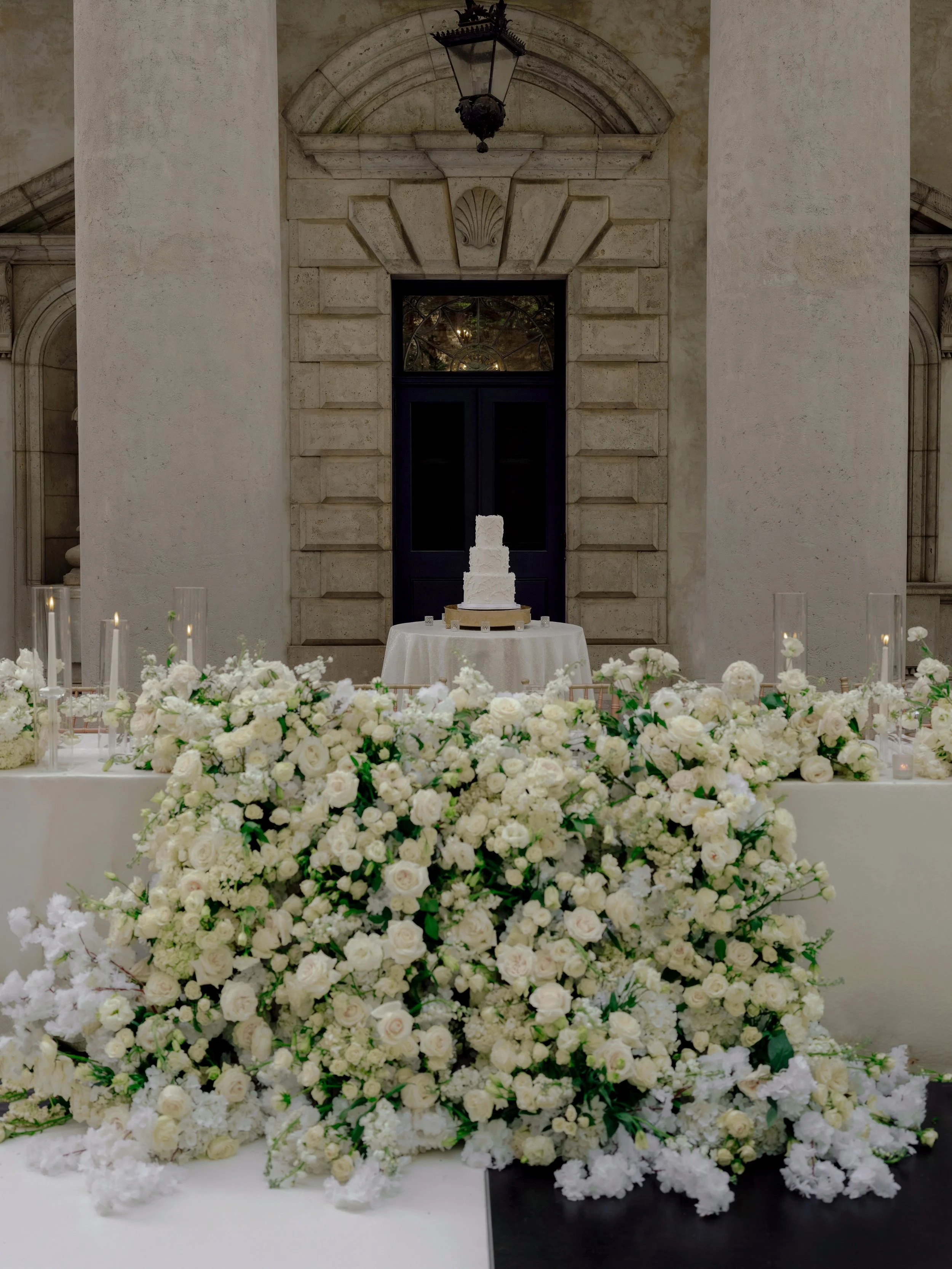 A wedding setup with a large floral arrangement of white roses and flowers in front of a table with a white wedding cake on it, against a historic stone building background.